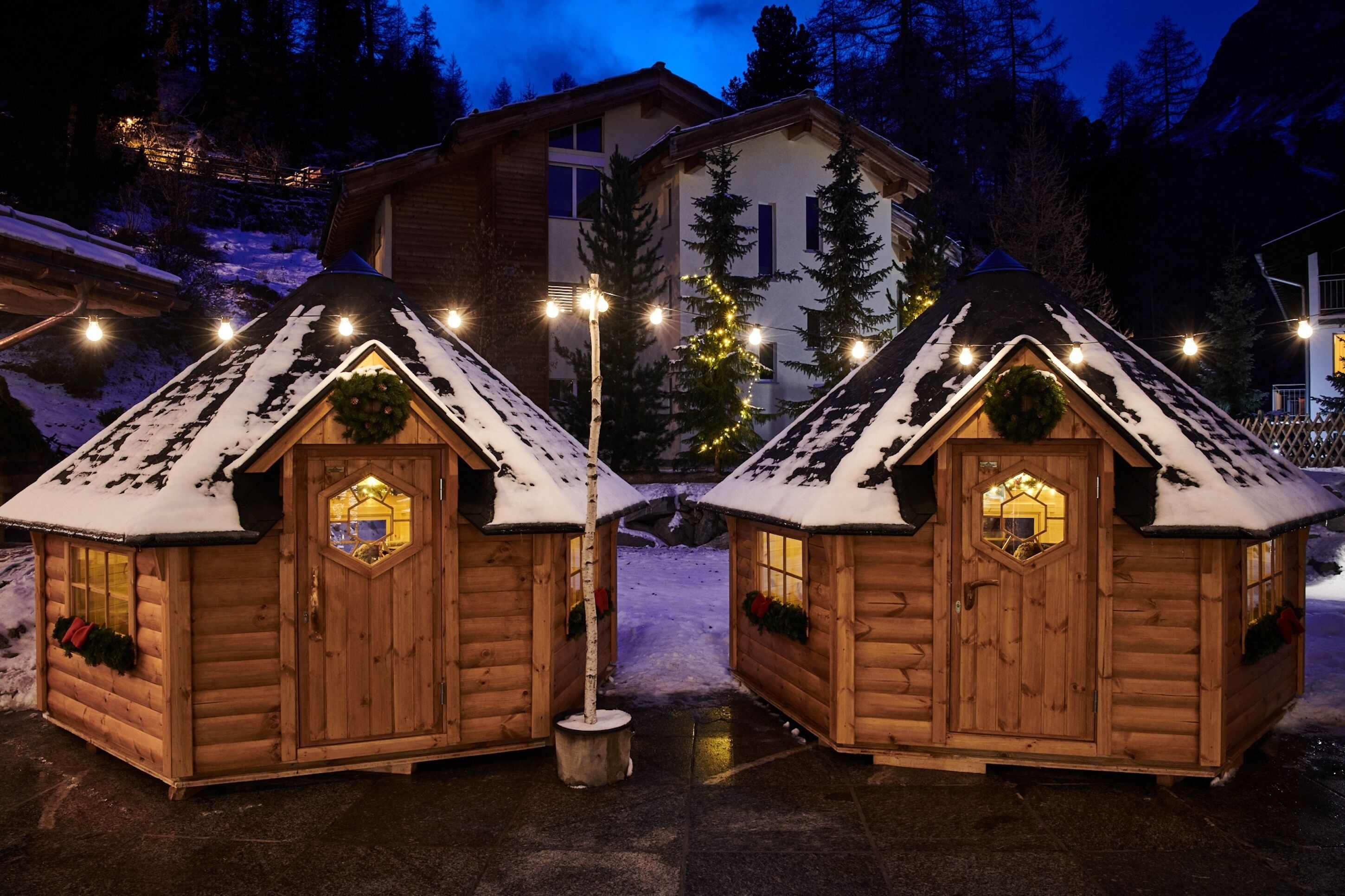 Two wooden cabins with snow-covered roofs, a Christmas wreath on each door, and a lit pathway leading to a larger building in the background.