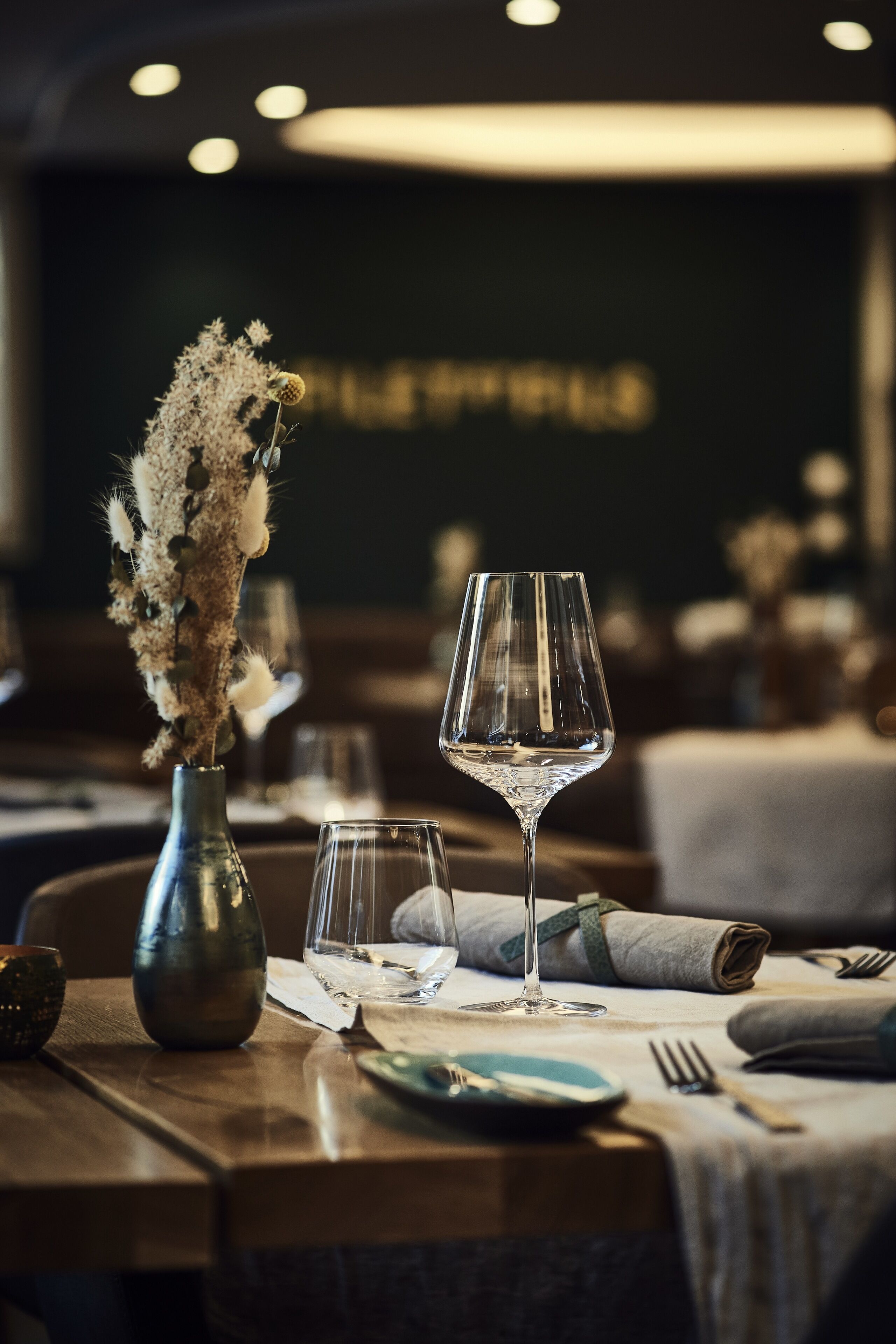 Una mesa de comedor con un jarrón de flores secas, una copa de vino y cubiertos dispuestos con cuidado.