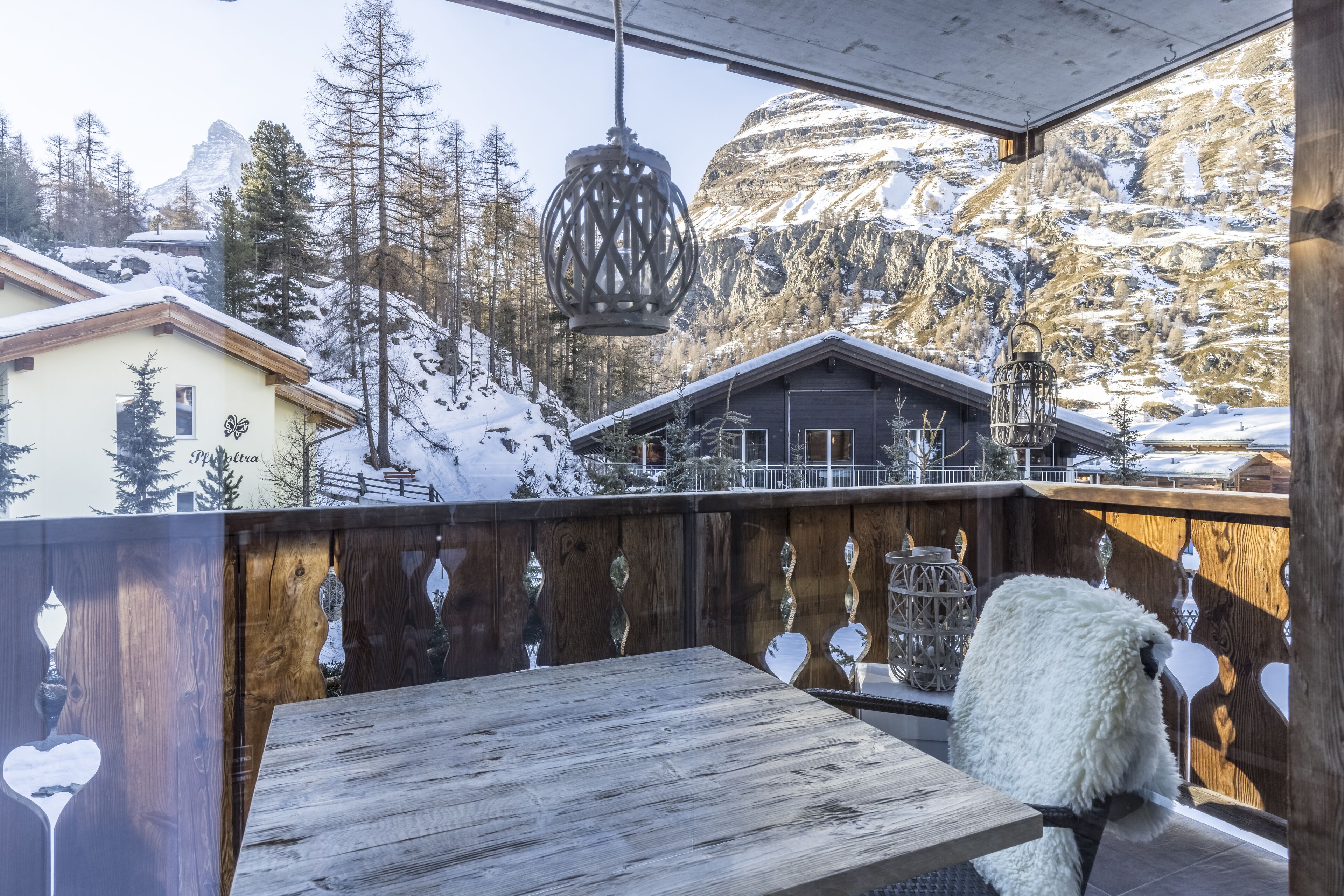 A wooden balcony with a table and chairs, overlooking a snowy mountain landscape with houses and trees.
