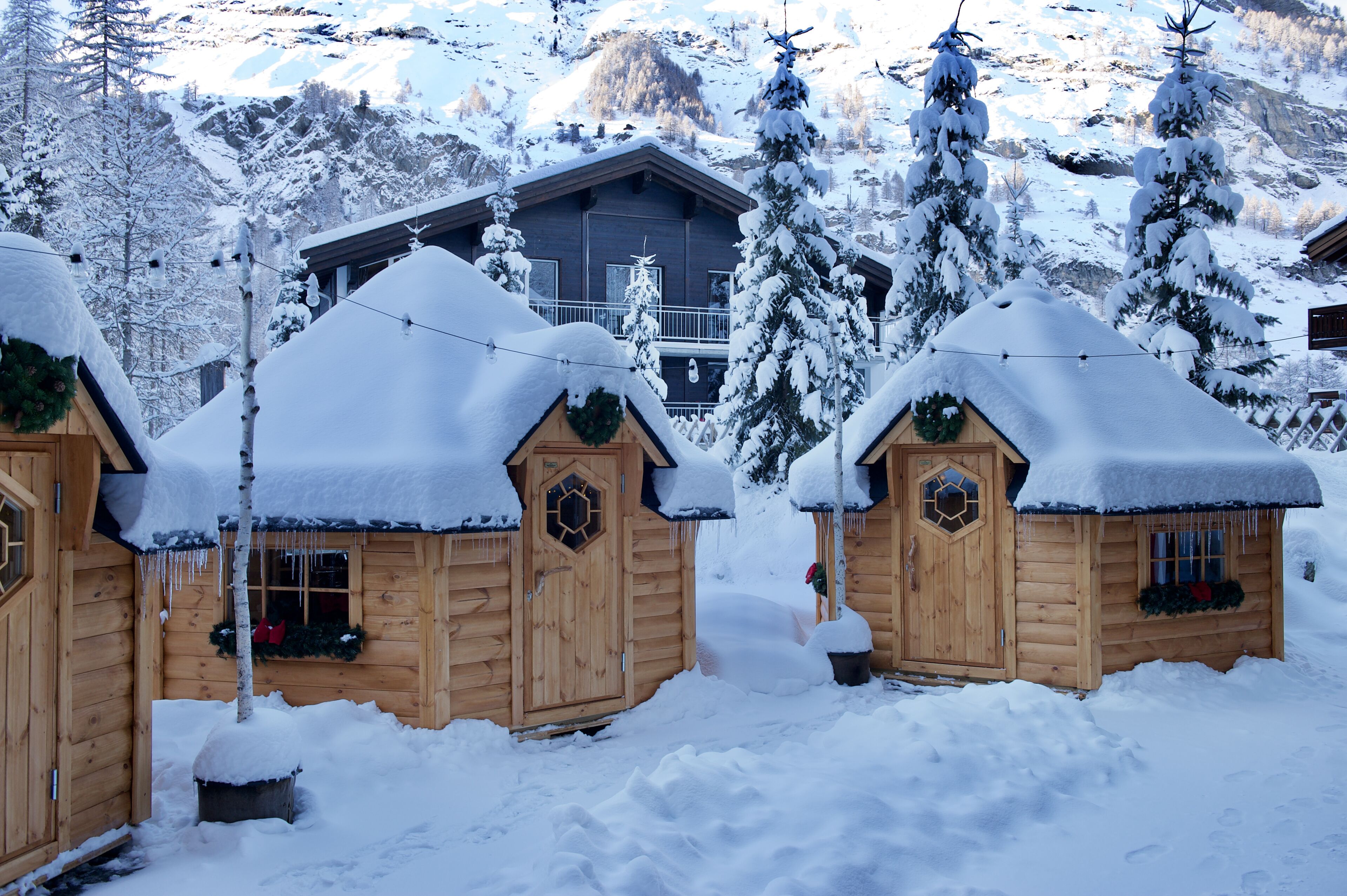 Una cabaña de madera con techo cubierto de nieve, rodeada de árboles nevados y montañas al fondo.