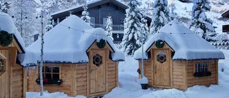 Una cabaña de madera con techo cubierto de nieve, rodeada de árboles nevados y montañas al fondo.