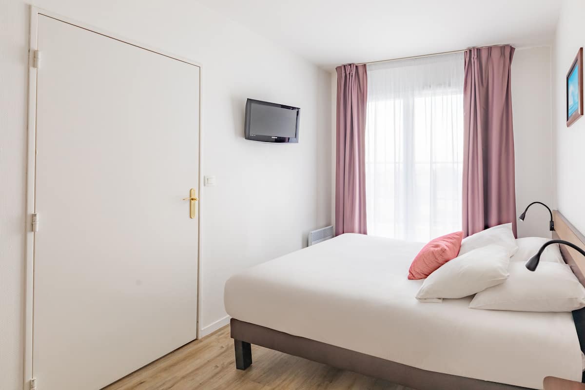 Una camera da letto con un letto, una TV a muro, una finestra con tende e un termosifone.