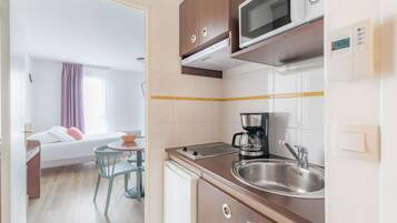Een compacte hotelkamer met kitchenette, een bed met kussens, een kleine eettafel en een raam met gordijnen.