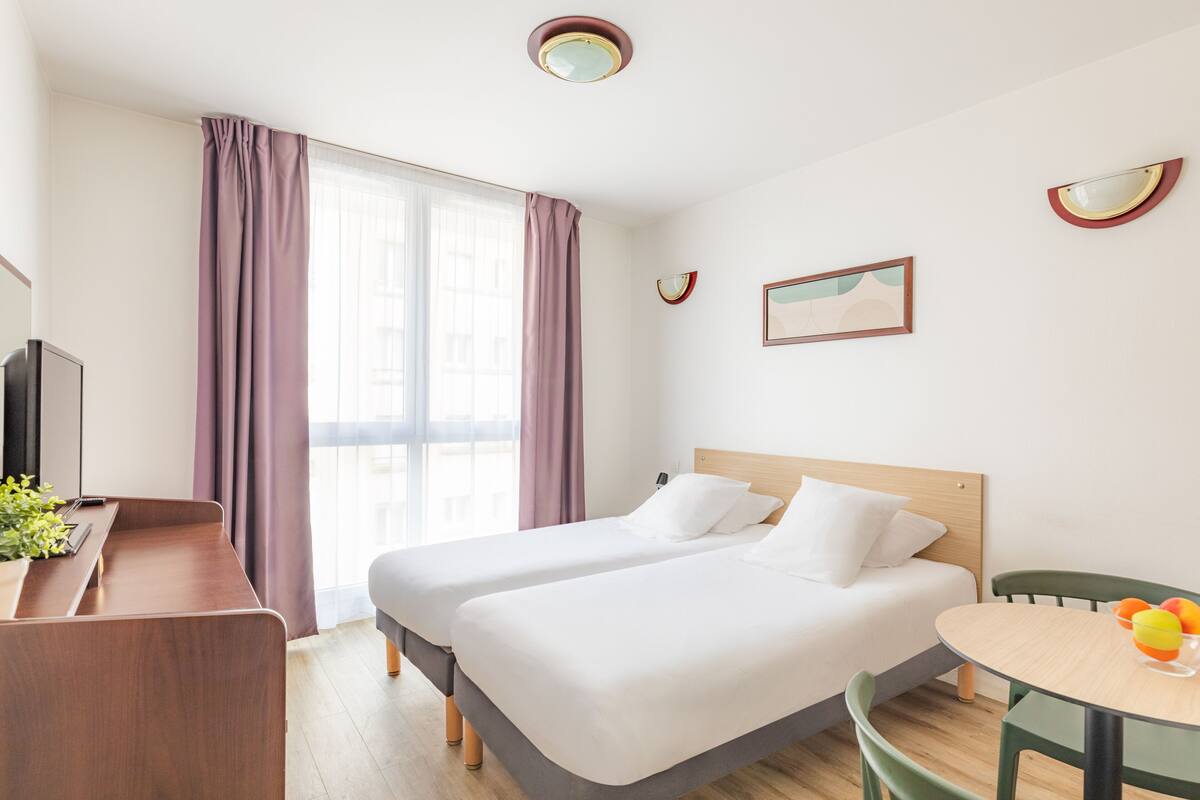 Camera d'albergo con due letti, una scrivania, una sedia, un tavolo con della frutta e una televisione.