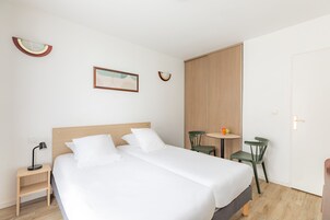 A hotel room with a double bed, a bedside table with a lamp, a small round table with chairs, and a wardrobe.