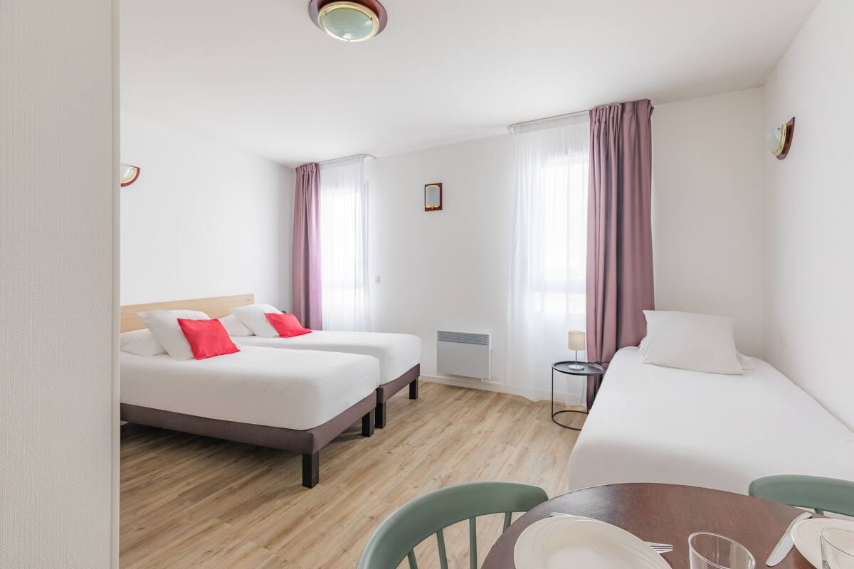 Una camera d'albergo con due letti singoli, un letto matrimoniale, un tavolo da pranzo con sedie e un comodino con una lampada.