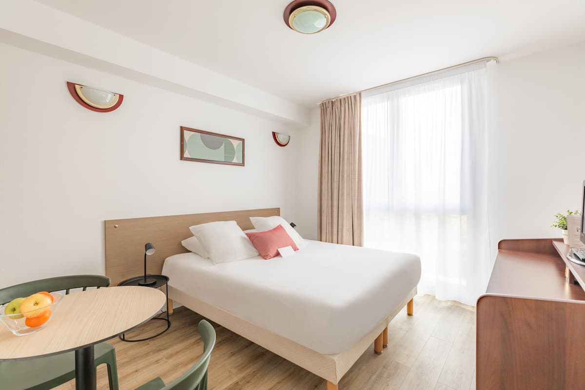Una moderna camera d'albergo con un letto, una scrivania, una sedia, un tavolino con della frutta e una finestra con tende.