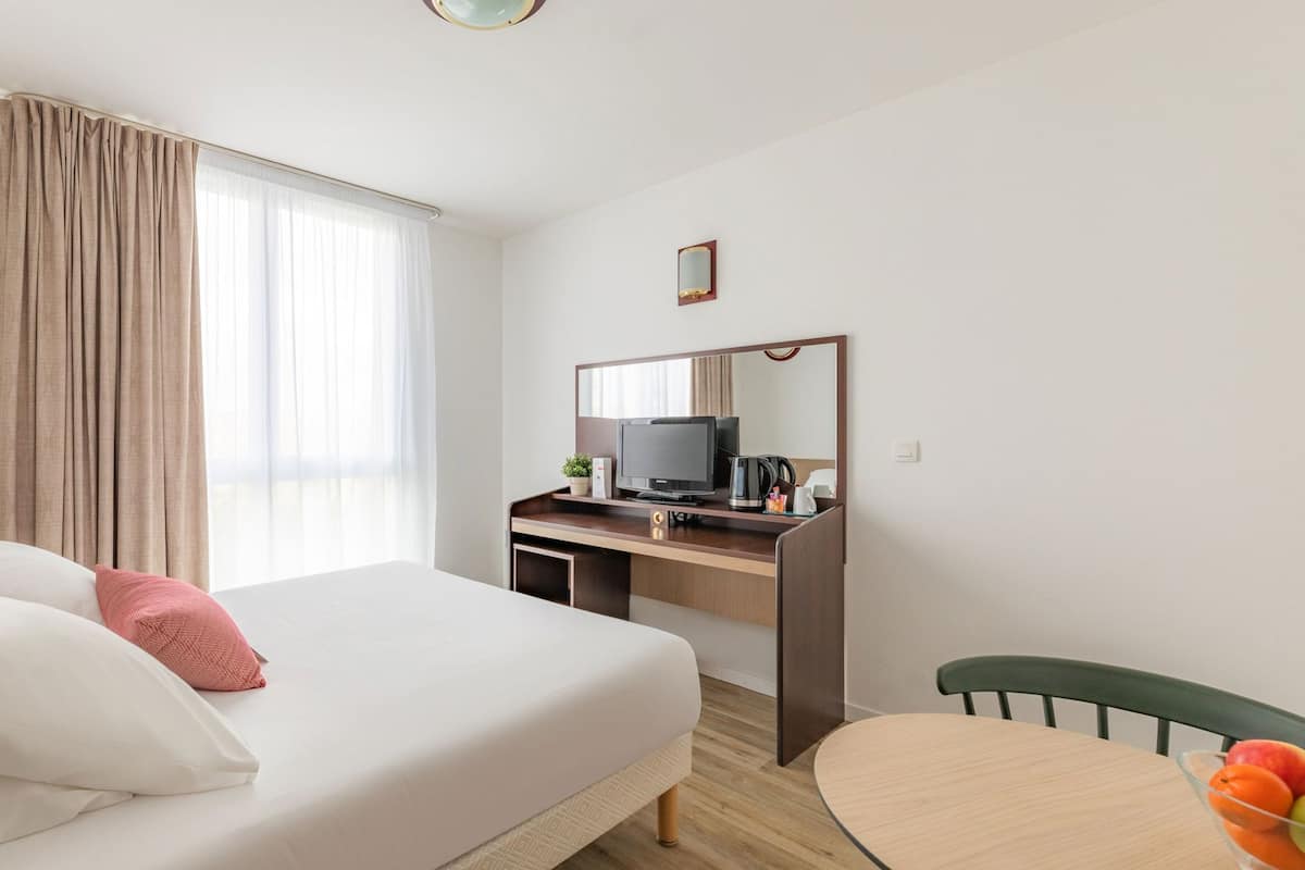 A hotel room with a bed, a desk with a TV, a chair, and a small table with fruit.