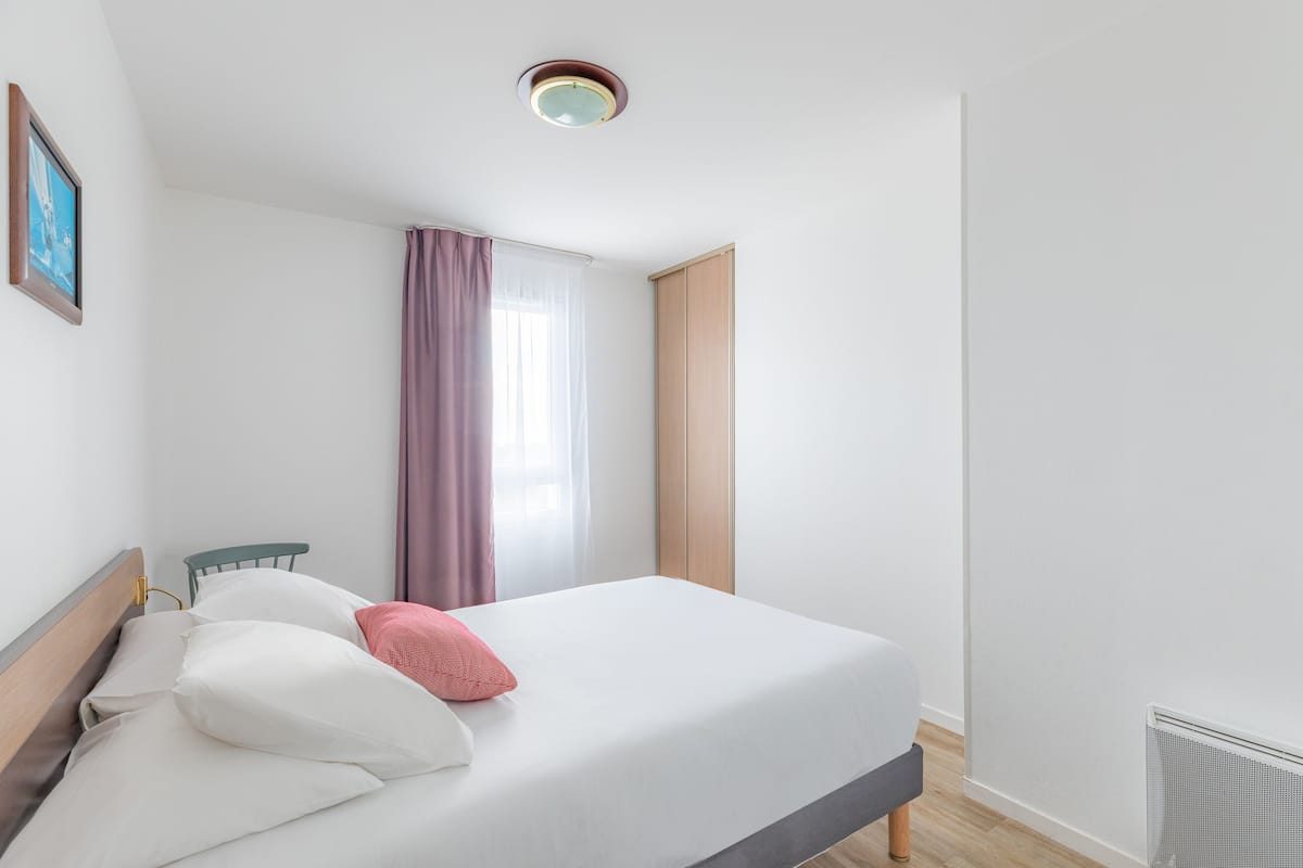 Un letto rifatto con cuscini bianchi e rosa, una testiera in legno e una finestra con tende viola.