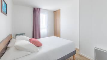 A neatly made bed with white and pink pillows, a wooden headboard, and a window with purple curtains.