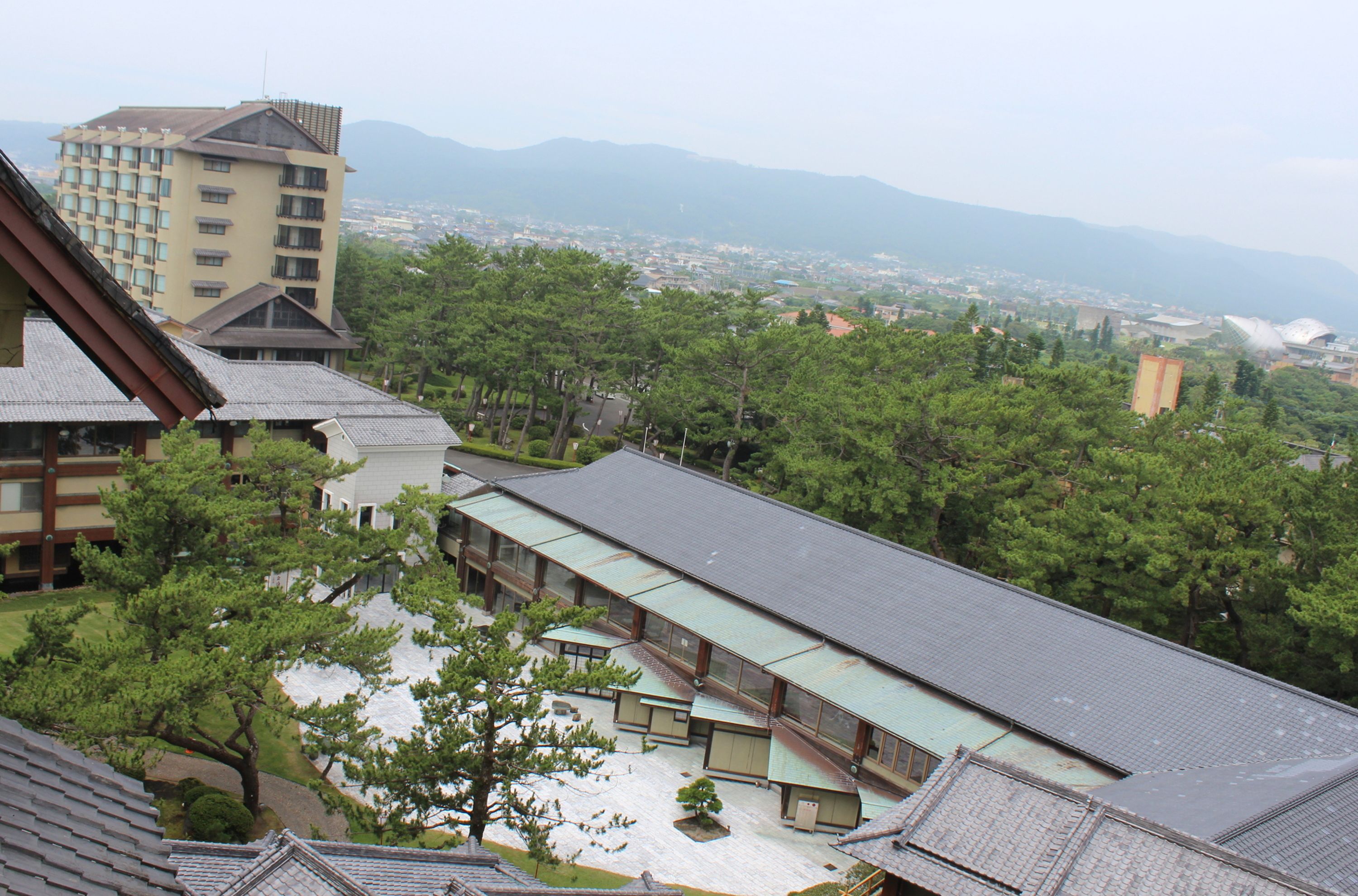 A traditional Japanese building with a sloped roof, surrounded by lush greenery and a view of distant mountains.