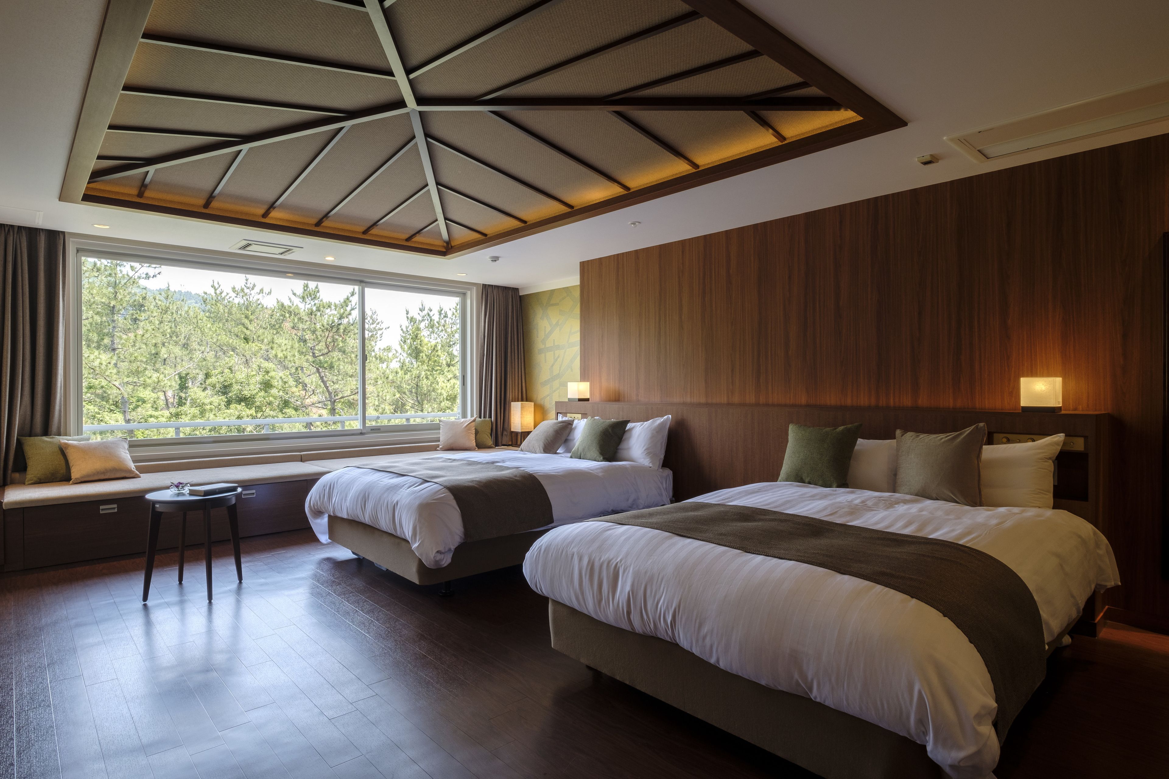 A modern hotel room with two beds, a large window, and a view of trees.