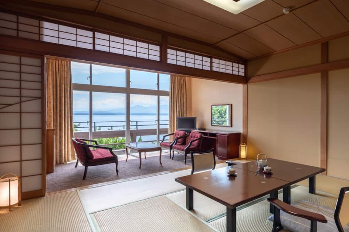 A traditional Japanese room with tatami flooring, a low table with cushions, a TV, and a view of the sea.