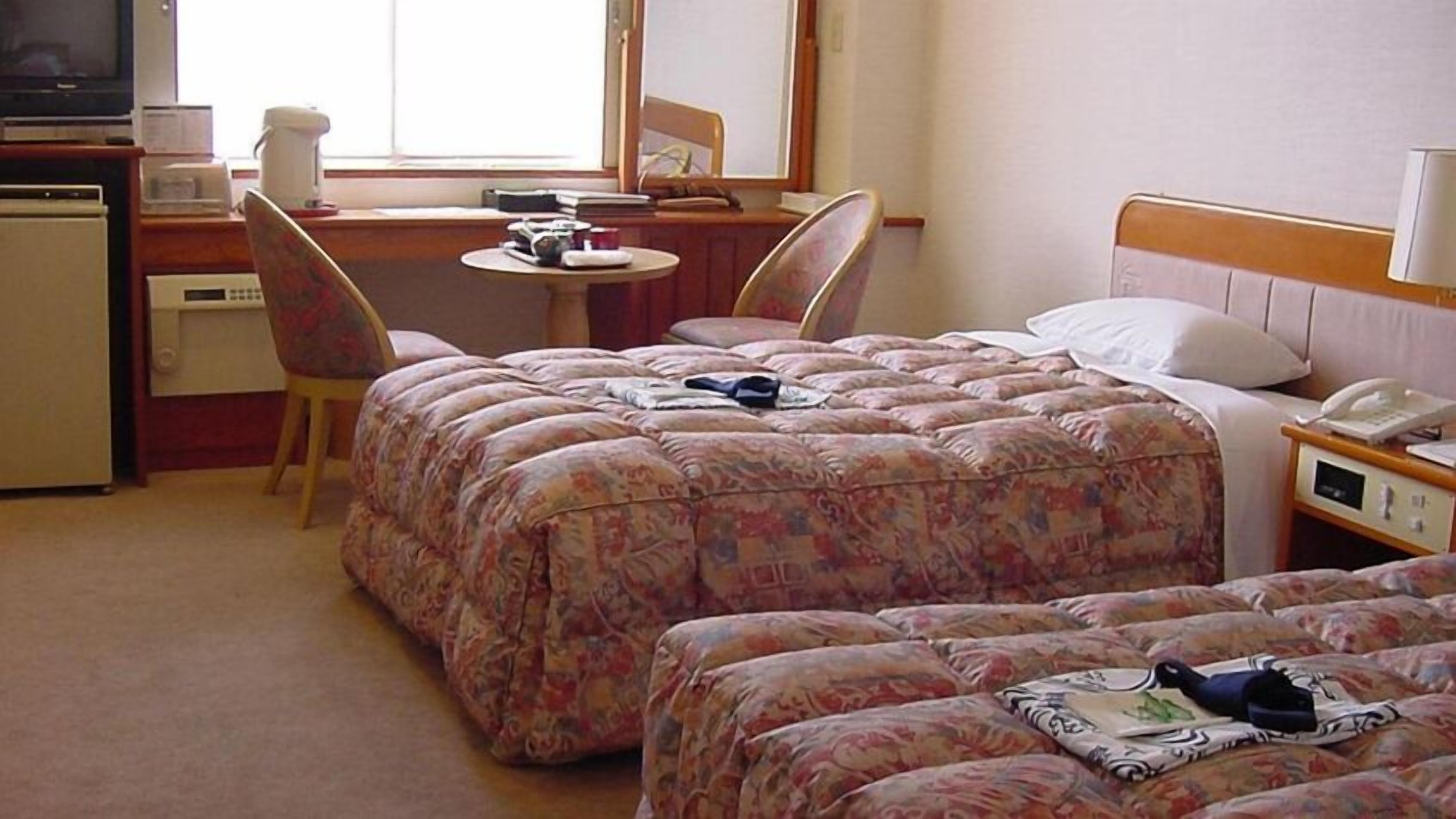 A hotel room with two beds, a desk, a chair, a small table, and a television.
