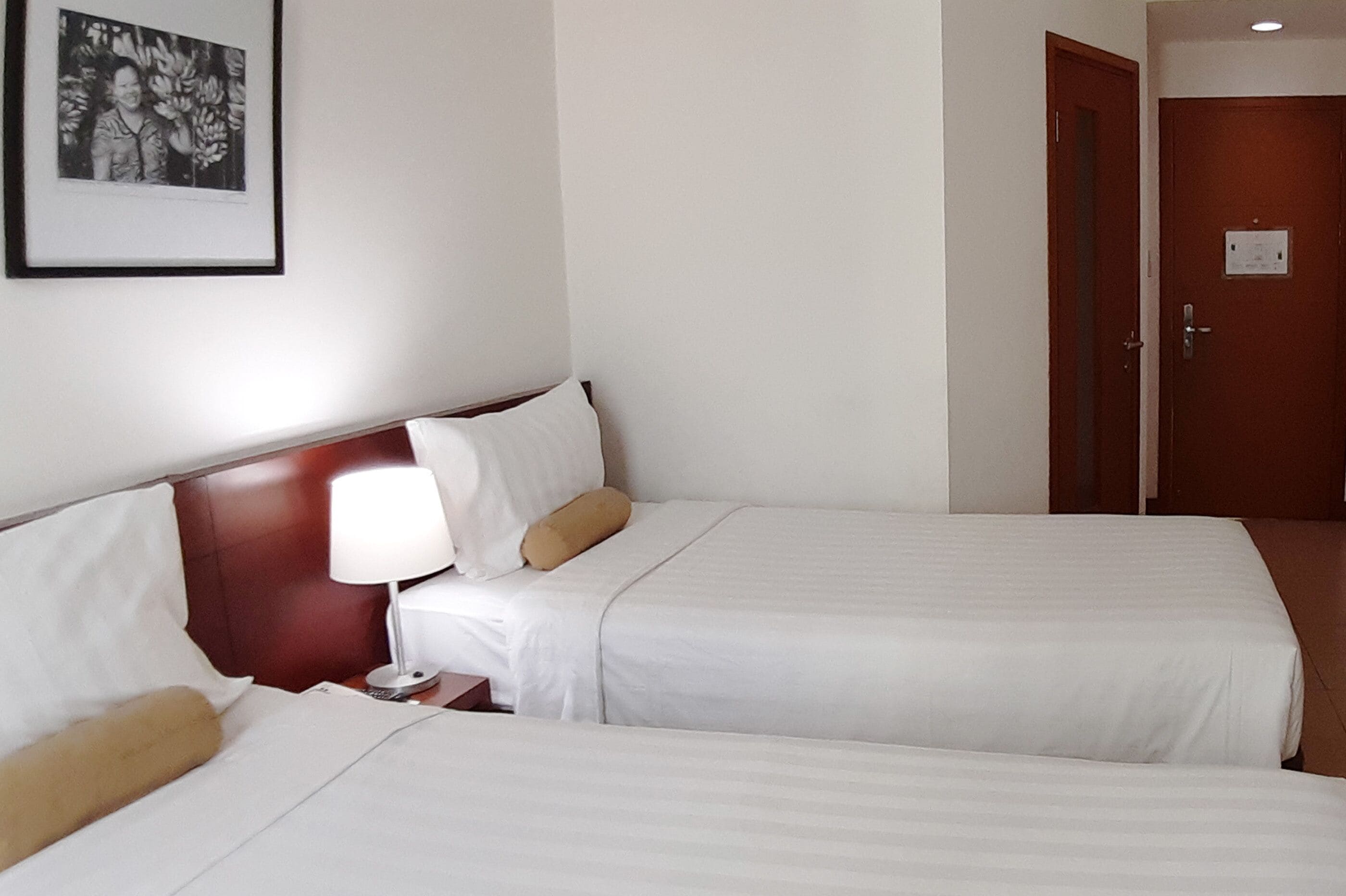 A hotel room with two beds, a nightstand with a lamp, a framed picture on the wall, and a door.