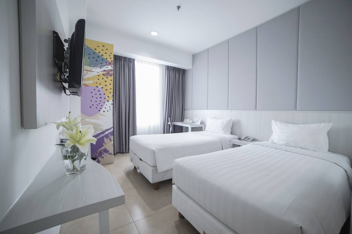 A hotel room with two beds, a small table with a vase of flowers, a TV mounted on the wall, and a window with curtains.