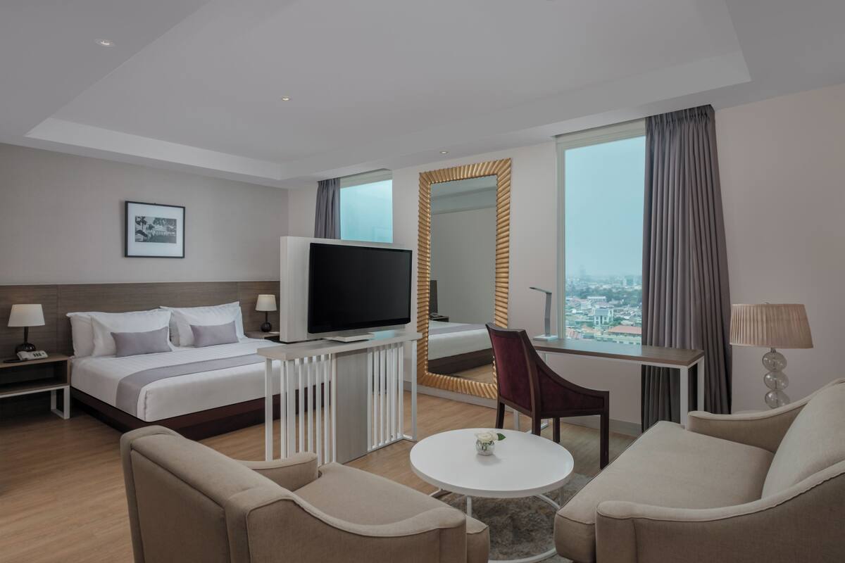 A hotel room with a bed, TV, desk, chair, and a view of the city.