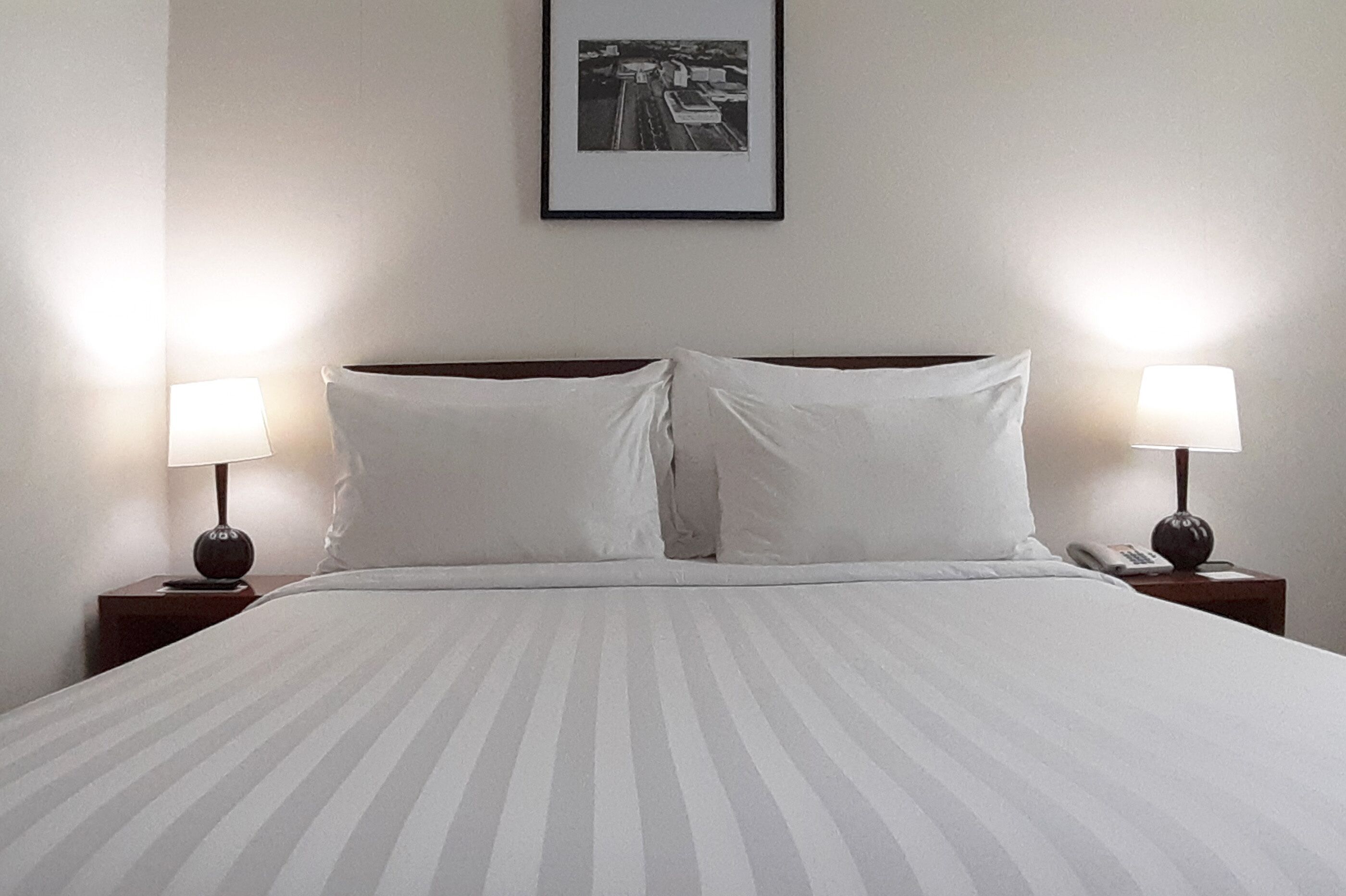 Een net opgemaakt bed met witte lakens, twee bedlampen en een ingelijste foto aan de muur.