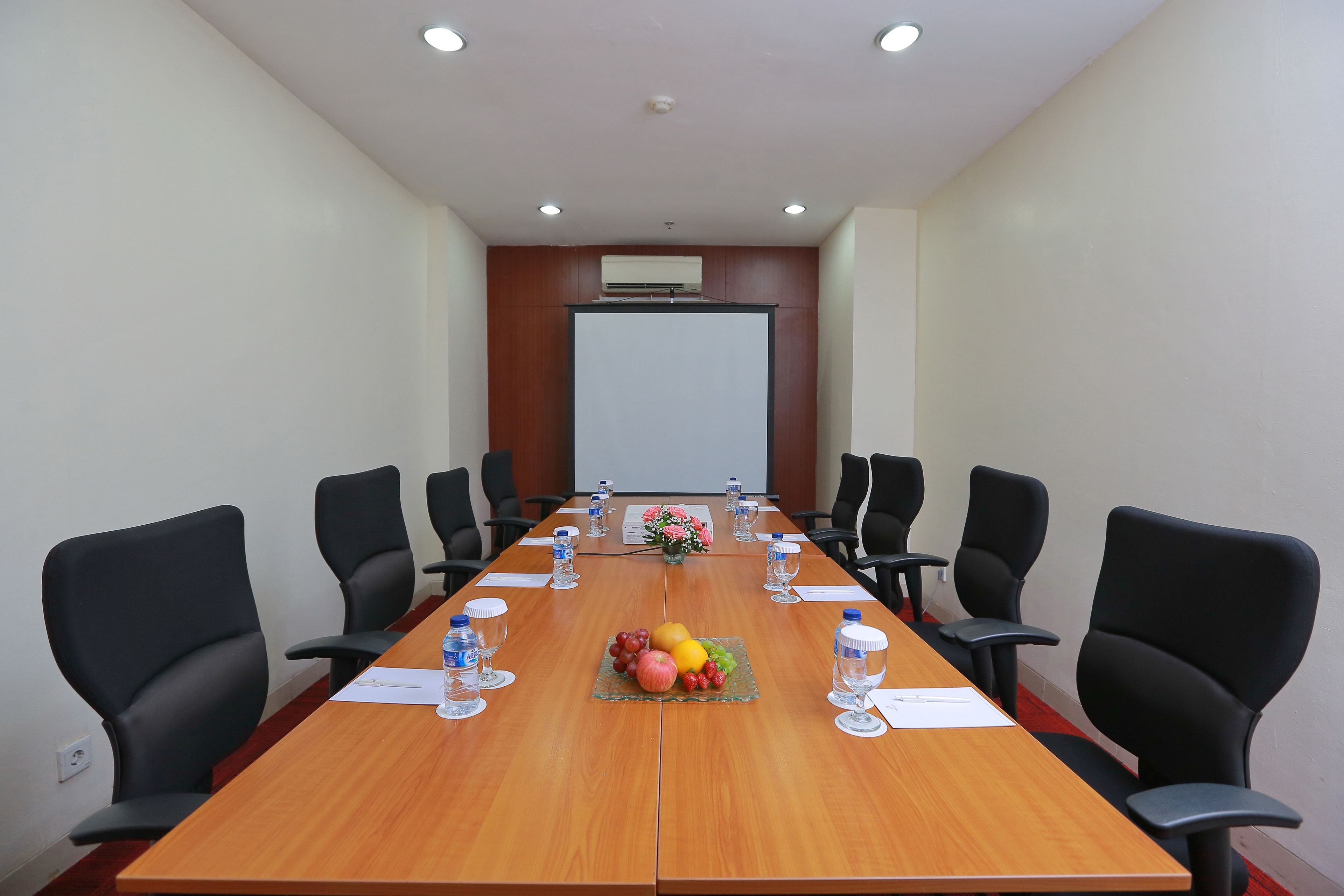 A conference room with a long wooden table, black chairs, water bottles, and a fruit basket.
