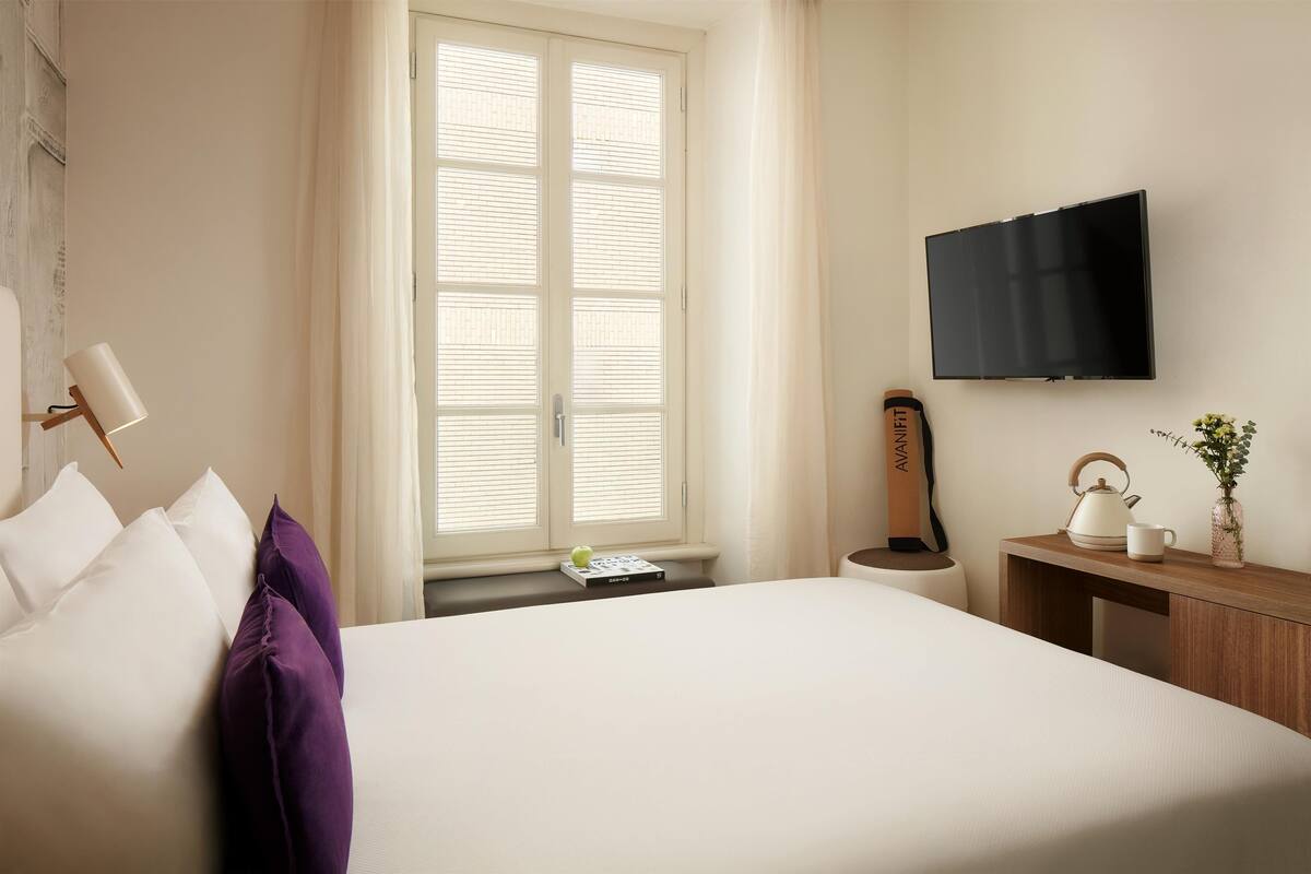 Une chambre d’hôtel avec un lit, un téléviseur fixé au mur, une fenêtre avec des stores, une table de chevet avec une bouilloire et un vase, et une lampe sur le côté gauche.
