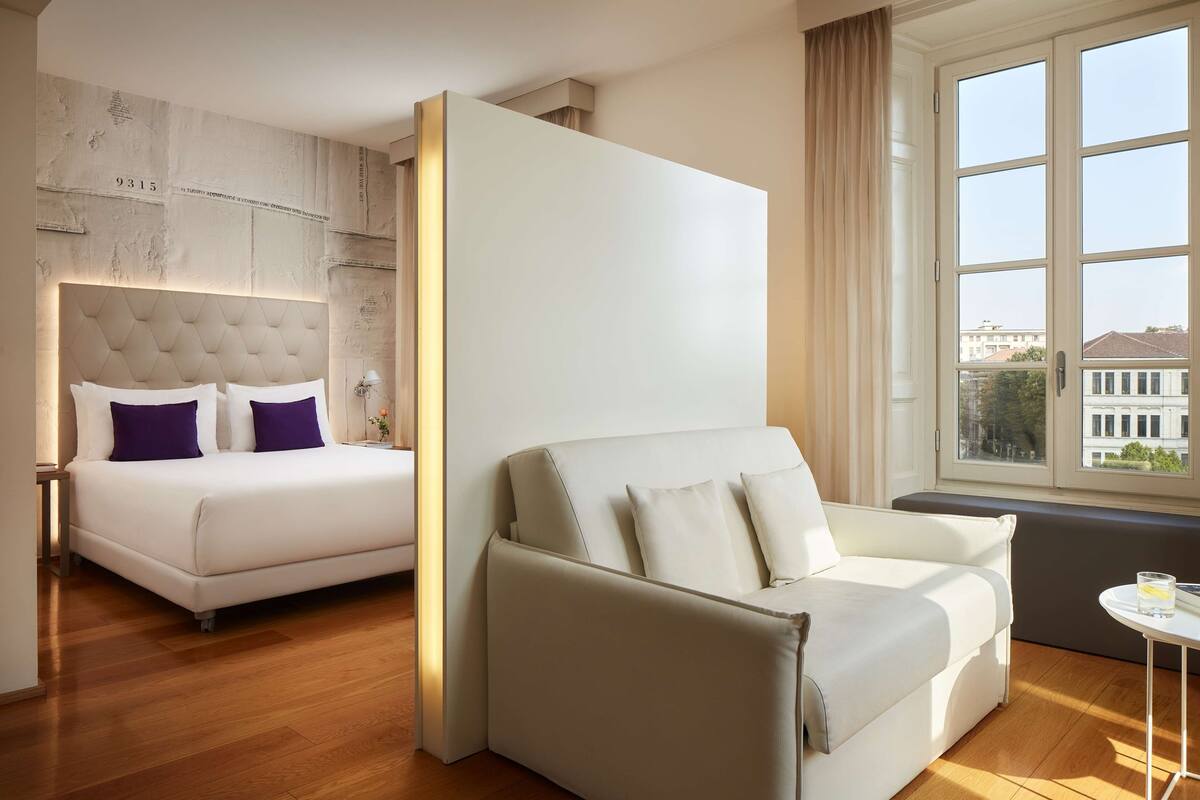 Une chambre d’hôtel avec un lit, un canapé, une petite table et une fenêtre donnant sur des immeubles.