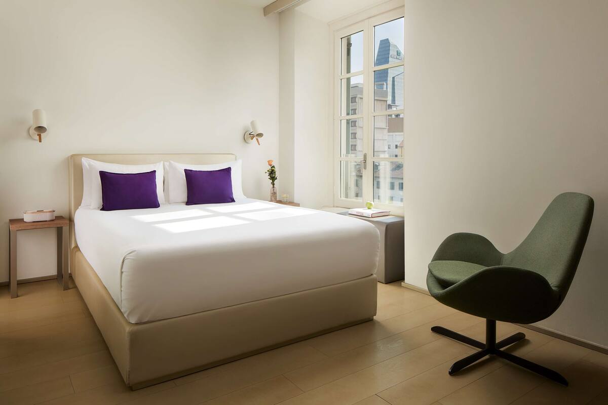 Une chambre avec un grand lit, un fauteuil vert, une table de chevet et une fenêtre donnant sur la ville.