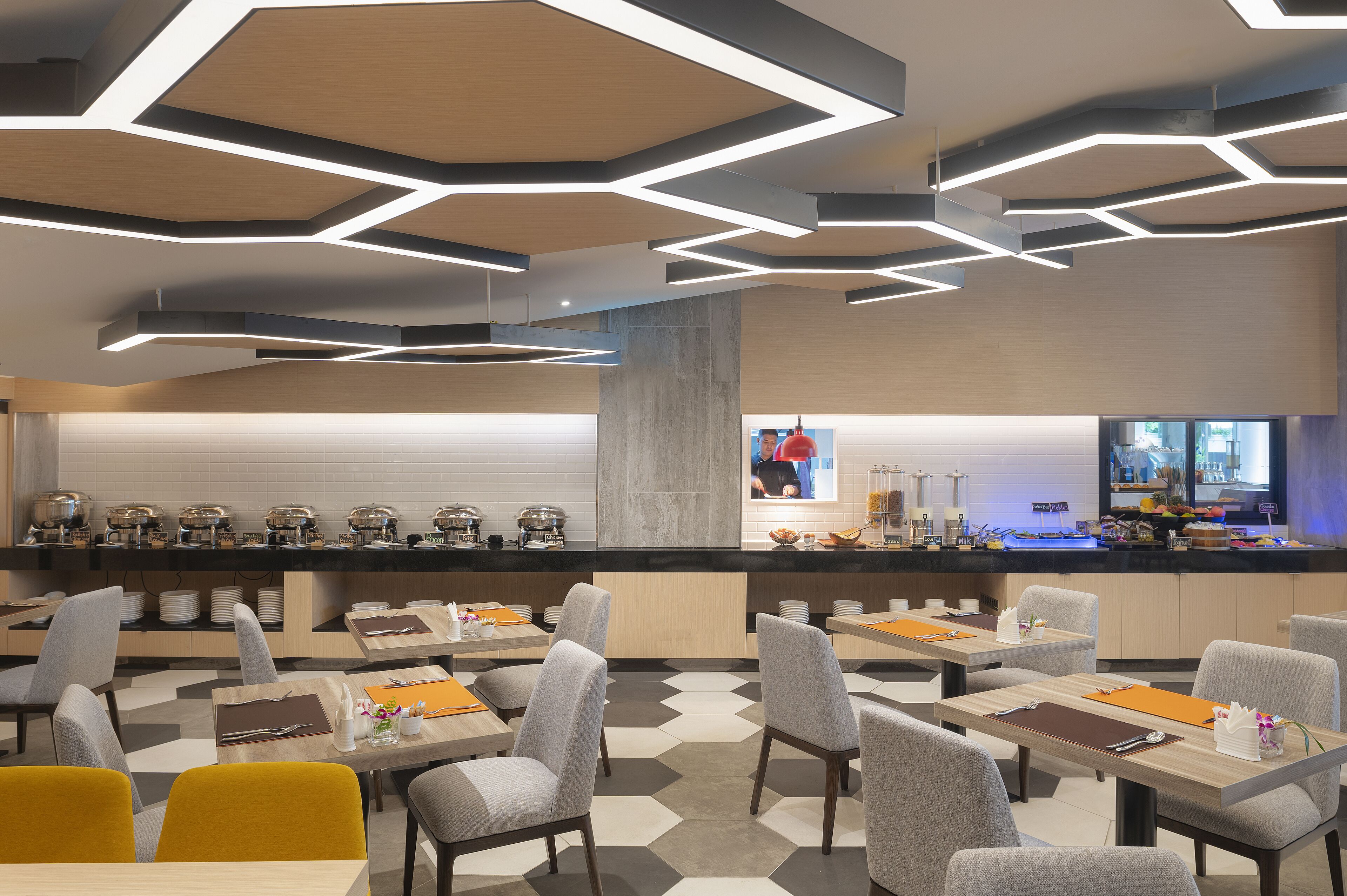 A modern restaurant with hexagonal ceiling lights, a counter with a variety of food items, and neatly arranged tables and chairs.