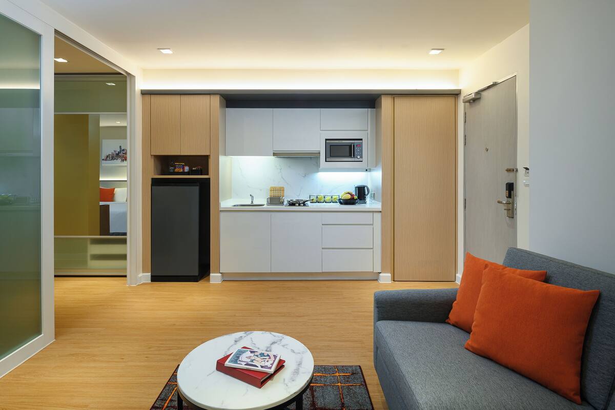 Un espacio habitable compacto con kitchenette, un sofá gris con cojines naranjas y una pequeña mesa redonda con un libro.