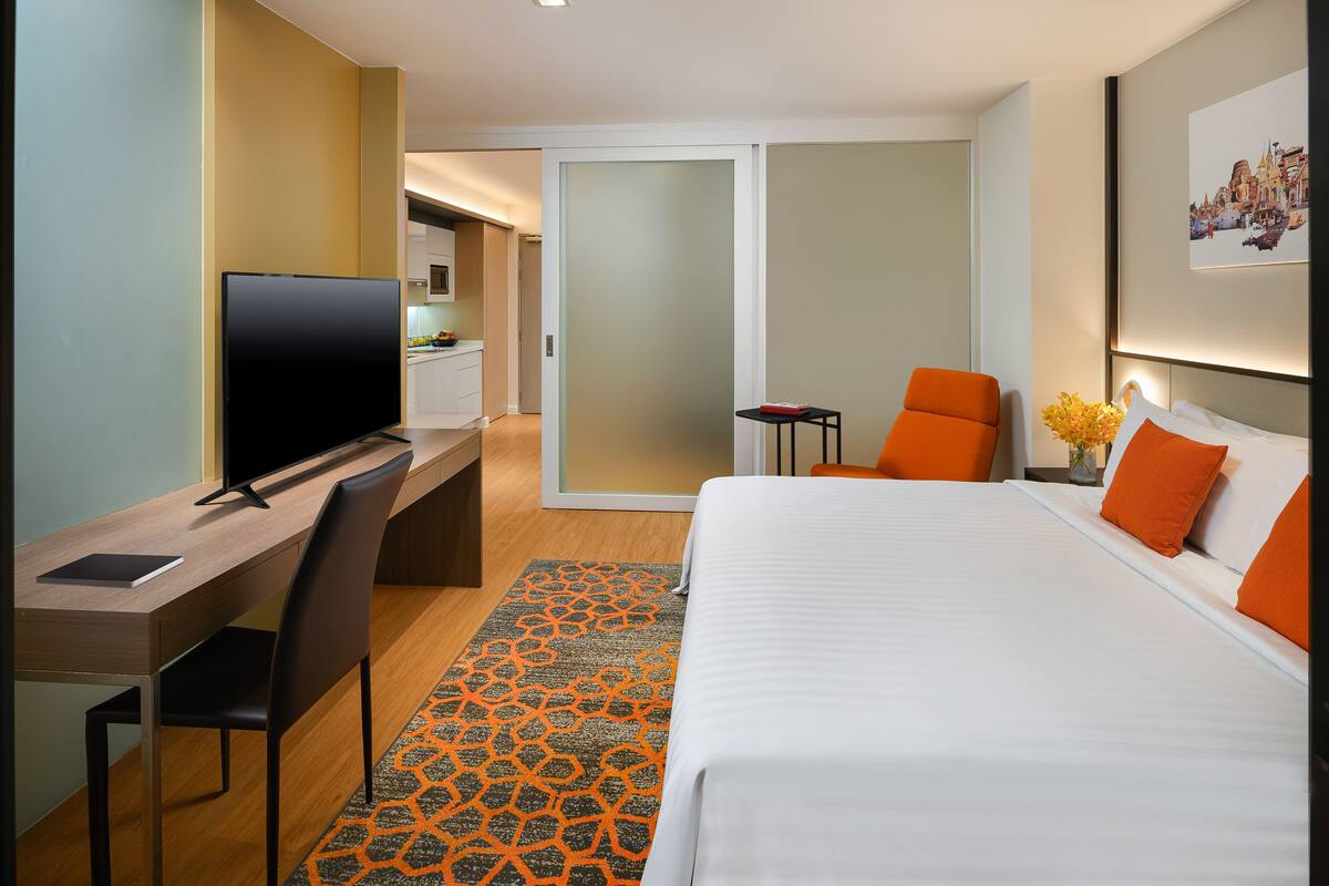 Habitación de hotel con cama, escritorio con televisor, silla, mesita pequeña y un jarrón decorativo.