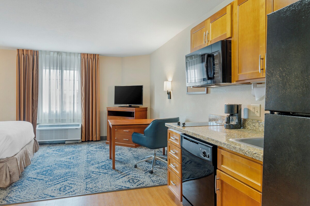 Habitación de hotel con cama, escritorio con silla, televisión, microondas, refrigerador y ventana con cortinas.