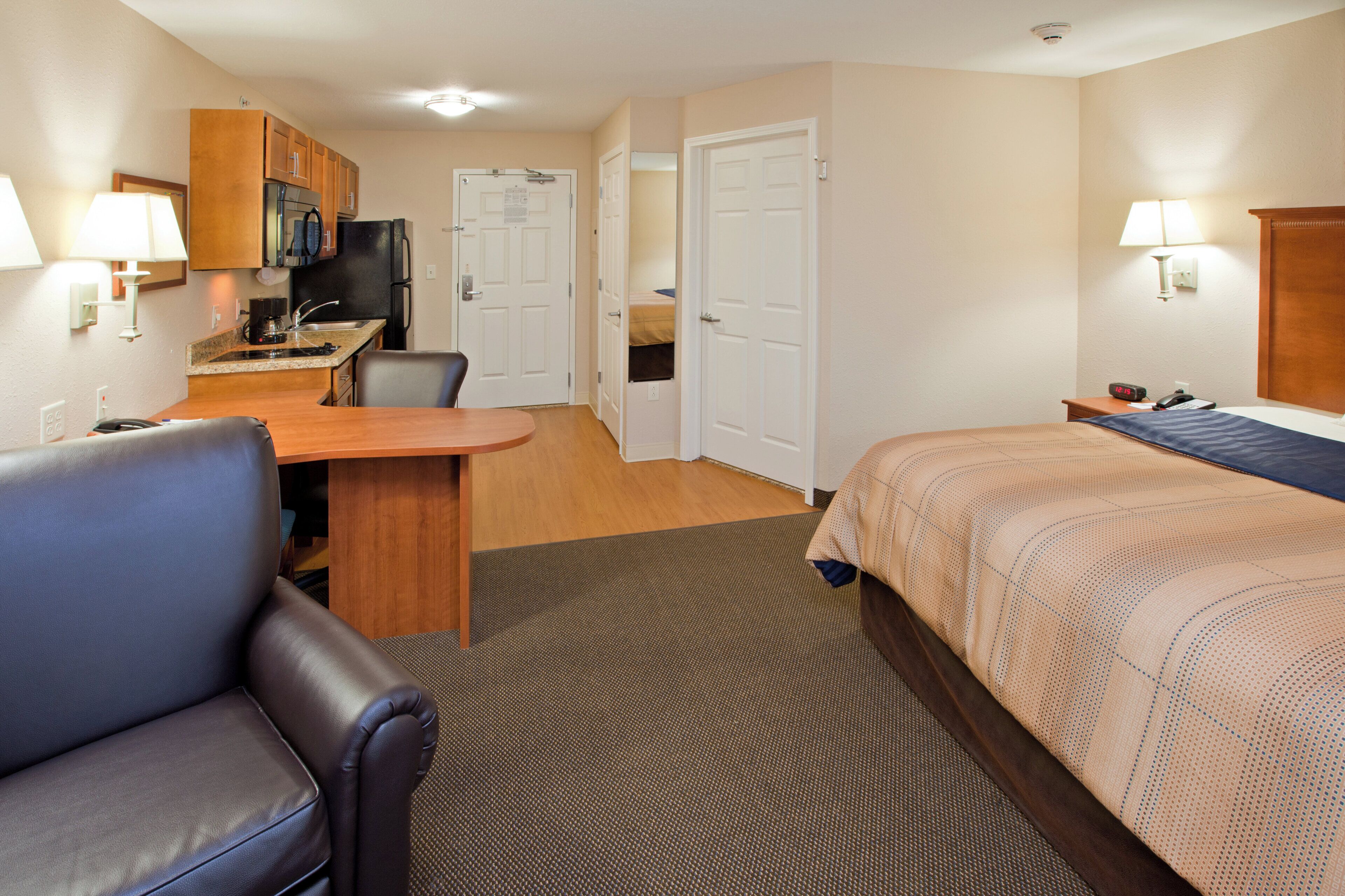 A hotel room with a bed, a desk, a chair, a kitchenette, and a bathroom.