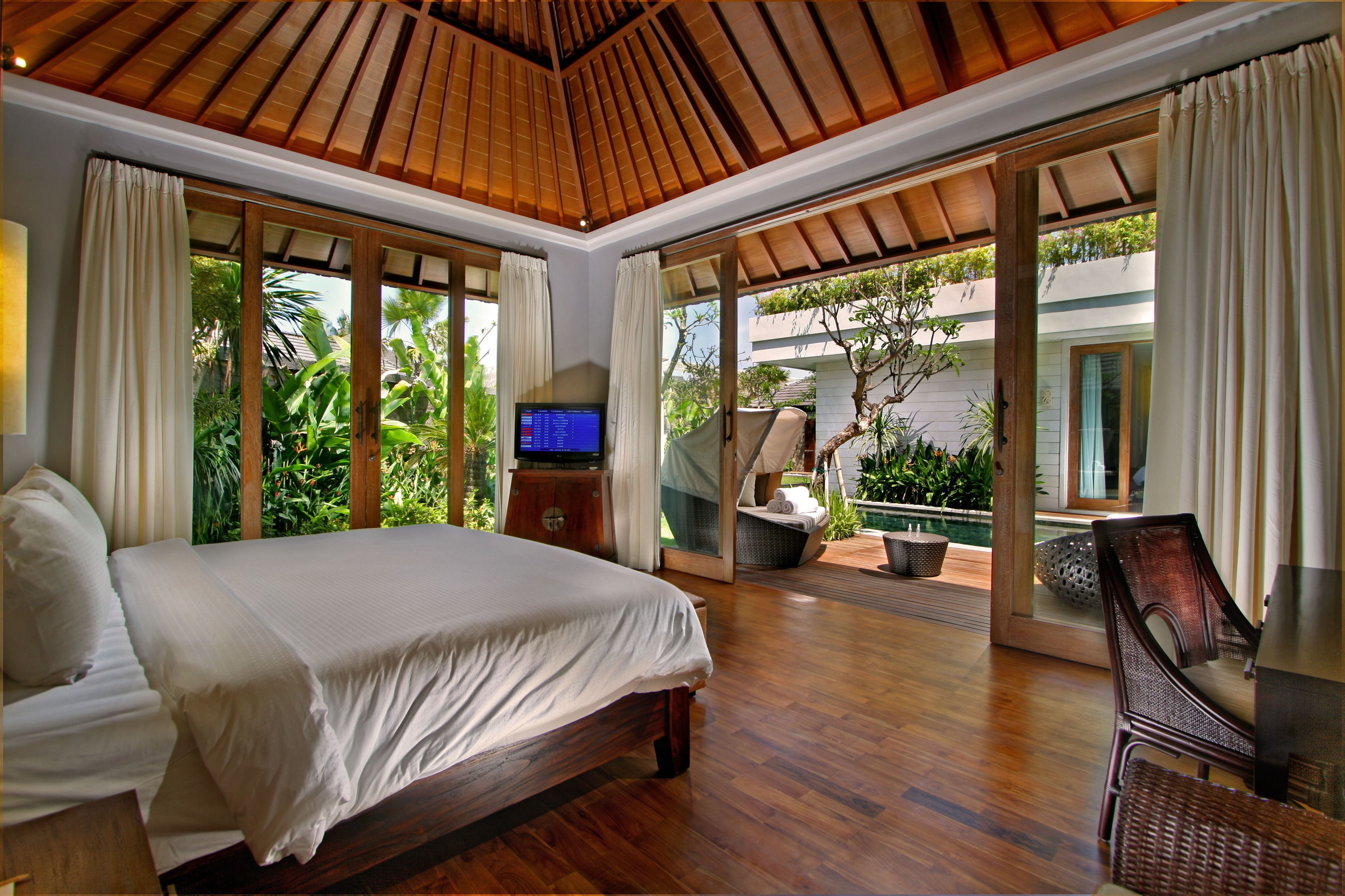 A spacious bedroom with a large bed, wooden flooring, a television, and a view of a pool and greenery.