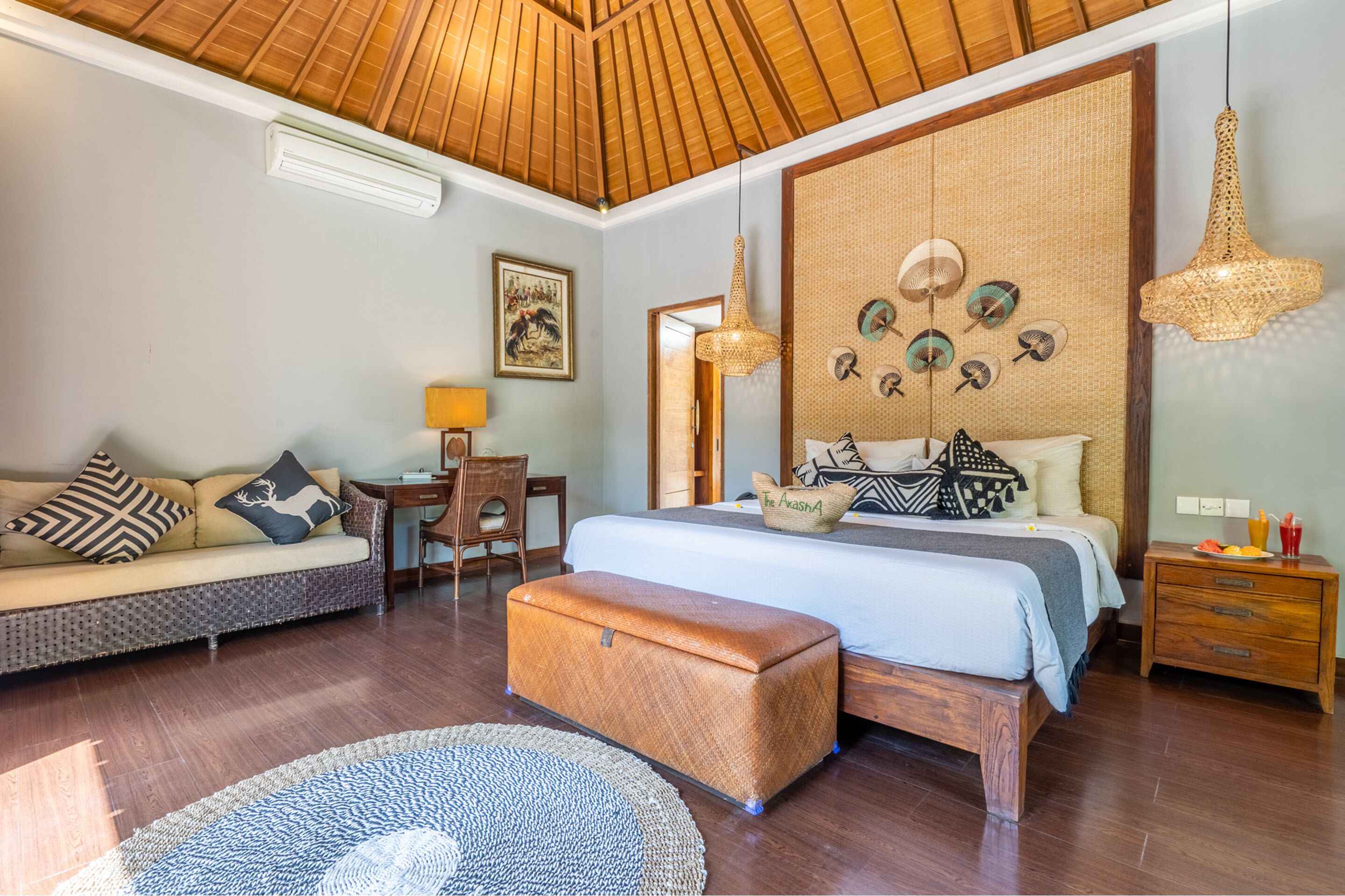 A well-lit bedroom with a wooden ceiling, a large bed with patterned pillows, a wooden bench, a desk with a lamp, and a wall decorated with circular objects.