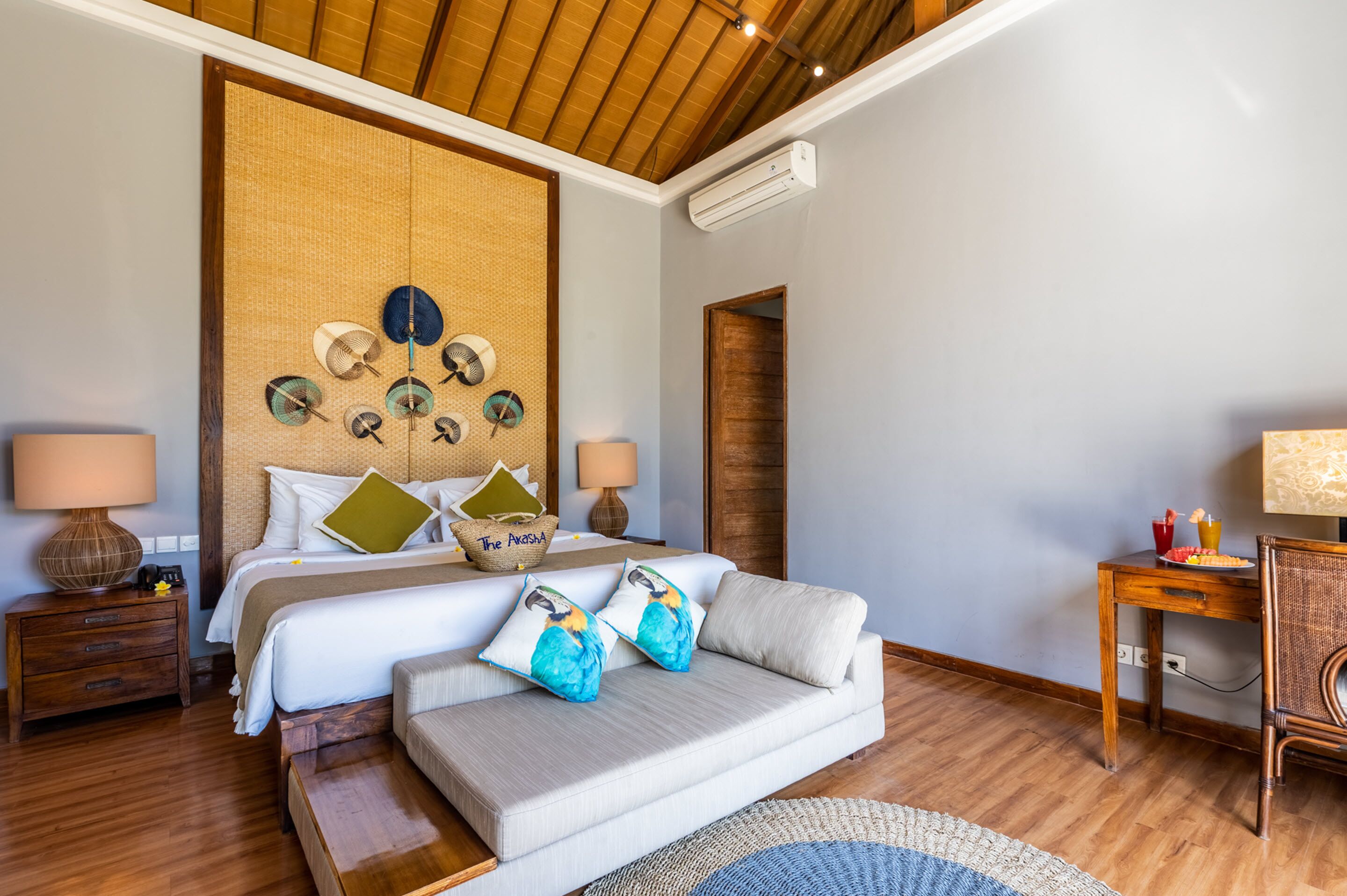 A hotel room with a large bed, a sofa, a wooden floor, a wall with decorative items, and a small table with a lamp.