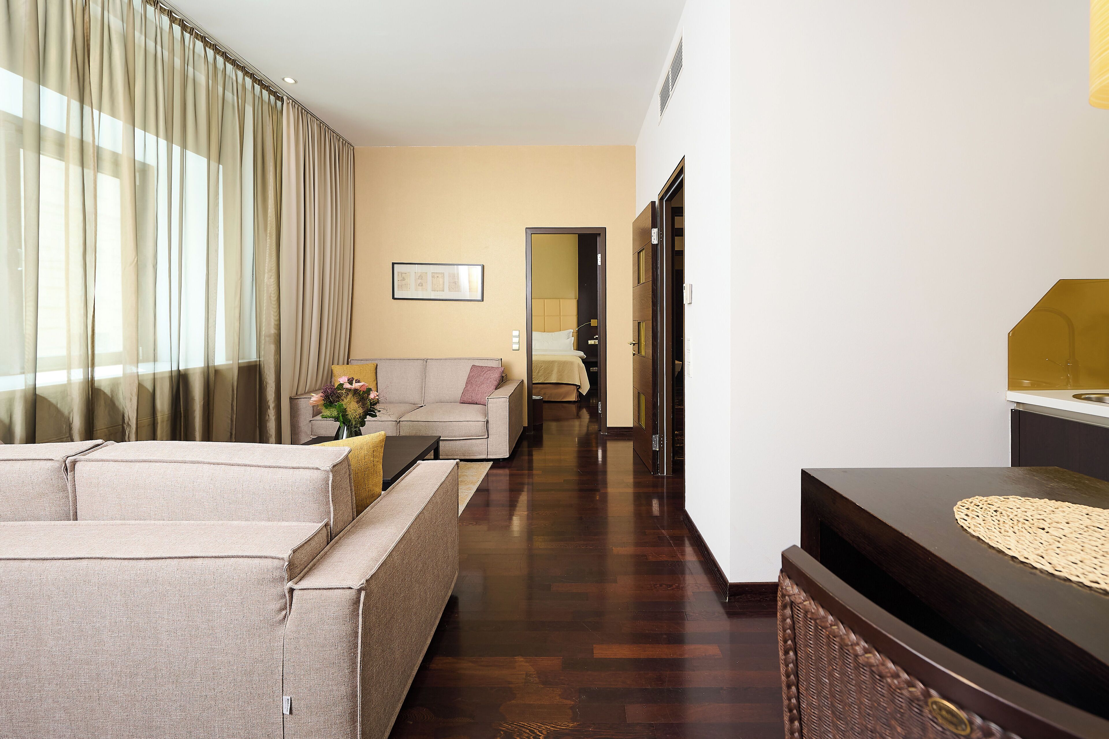 Quarto de hotel com sofá, uma mesinha com um vaso, um armário de madeira e uma cama ao fundo.