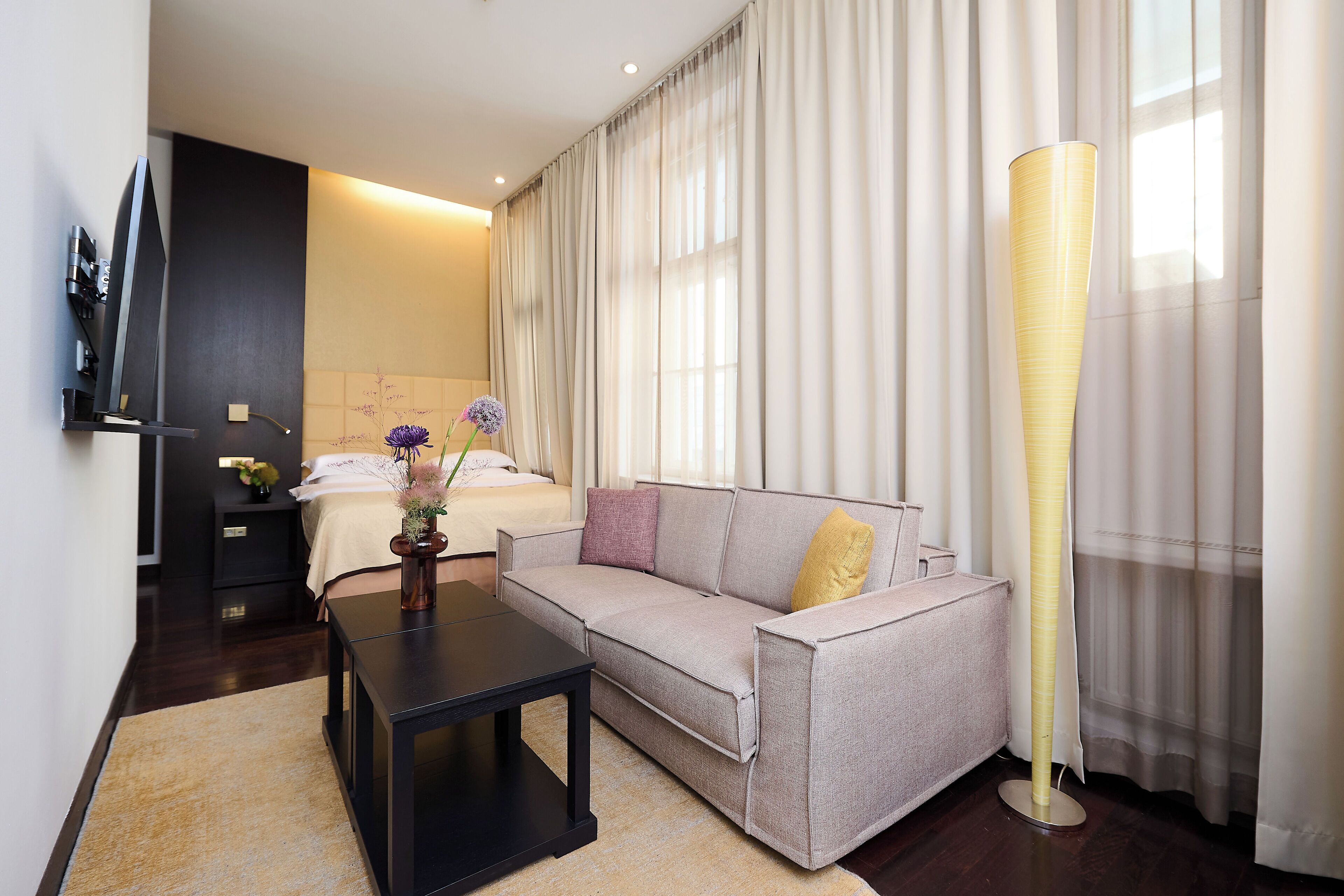 Quarto de hotel moderno com sofá, mesa de cabeceira, TV e uma janela grande com cortinas.