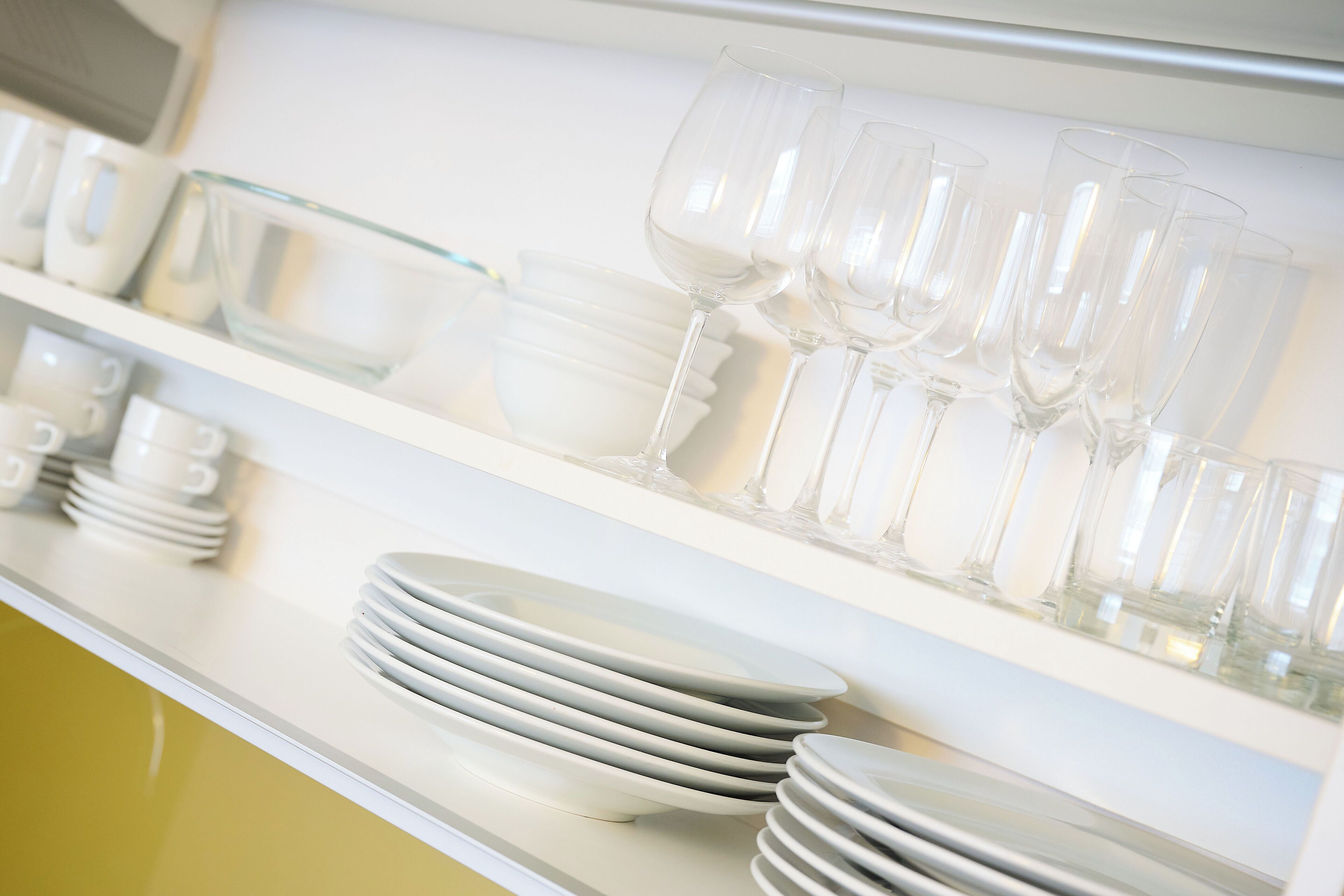 Une cuisine avec des meubles blancs et des étagères où sont empilées des assiettes blanches et des verres à vin transparents.