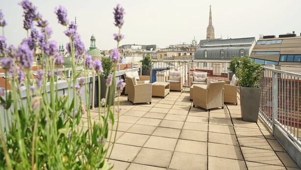 A rooftop terrace with wicker furniture, a potted plant, and a view of a city skyline including a church spire.