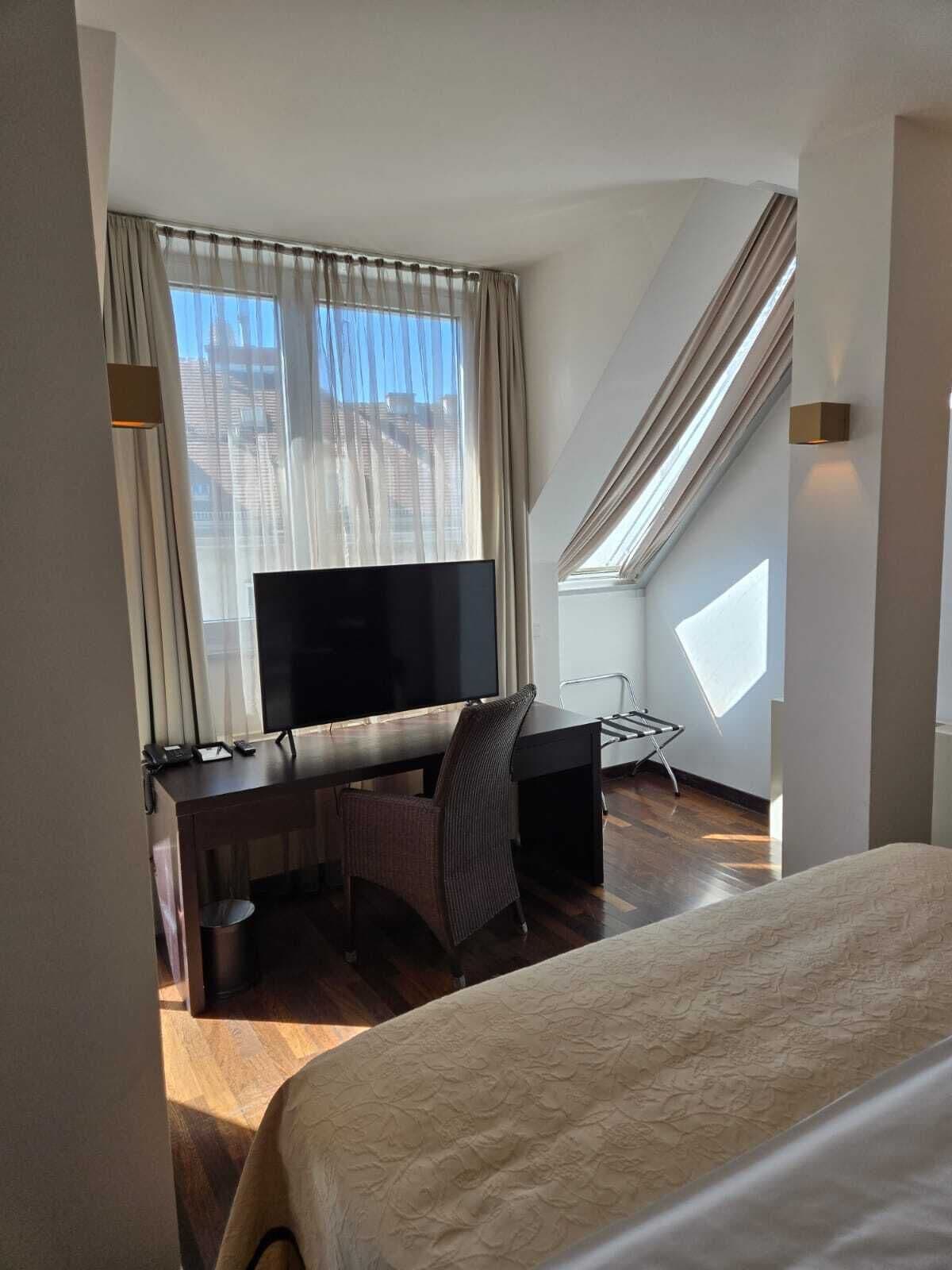 A hotel room with a bed, a desk with a TV, a chair, and a window with curtains.