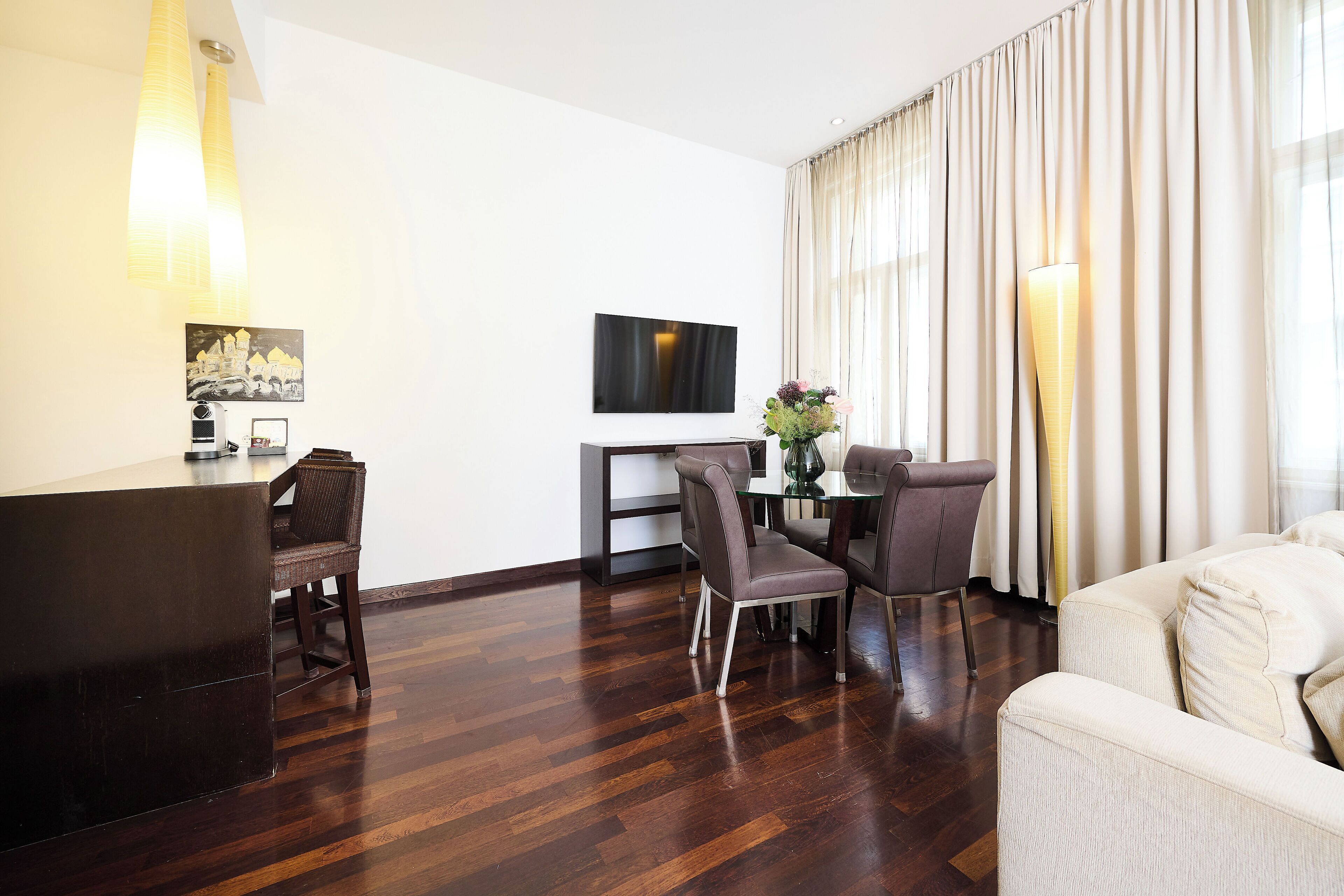 A modern hotel room with a wooden floor, a dining area with a glass table and chairs, a flat-screen TV mounted on the wall, and a large window with curtains.