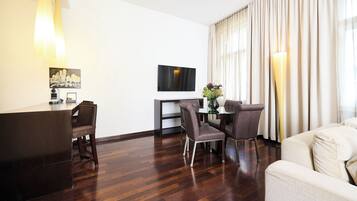 A modern hotel room with a wooden floor, a dining area with a glass table and chairs, a flat-screen TV mounted on the wall, and a large window with curtains.