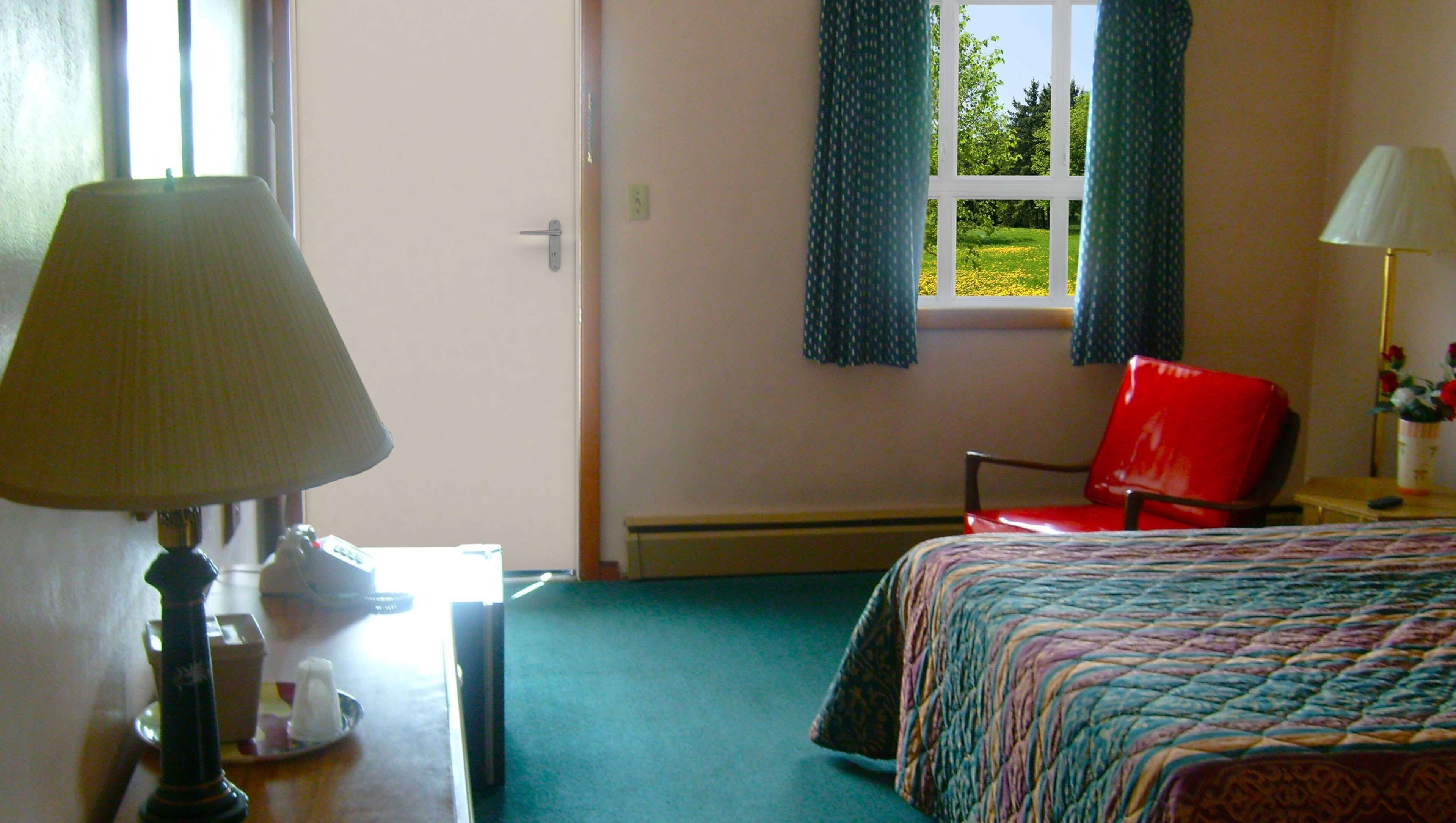 Habitación de hotel con cama, una silla, un escritorio con lámpara y ventana con cortinas.