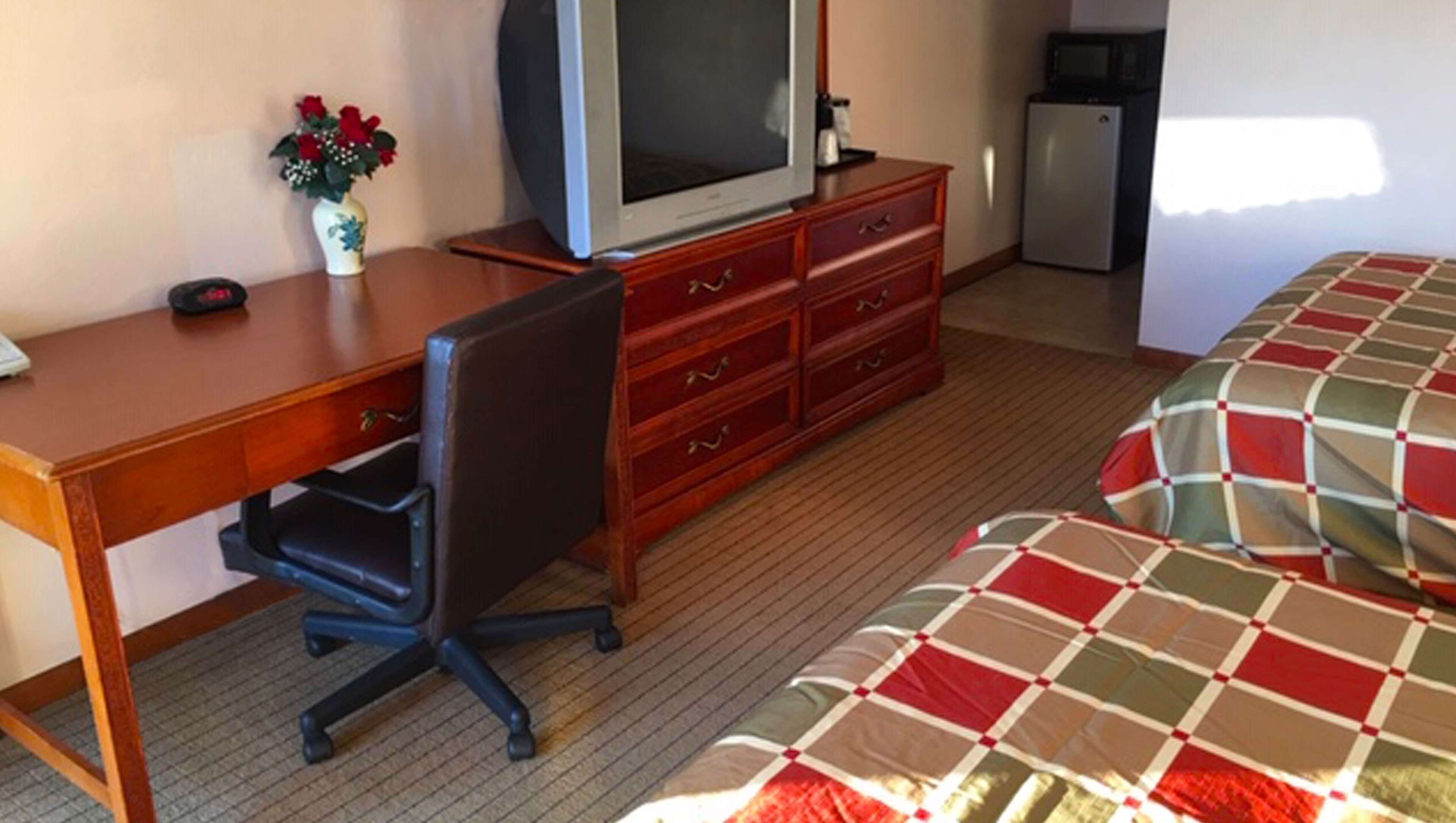 Hotelkamer met een houten bureau, een televisie, een dressoir, een kleine koelkast en een bed met een geruit sprei.