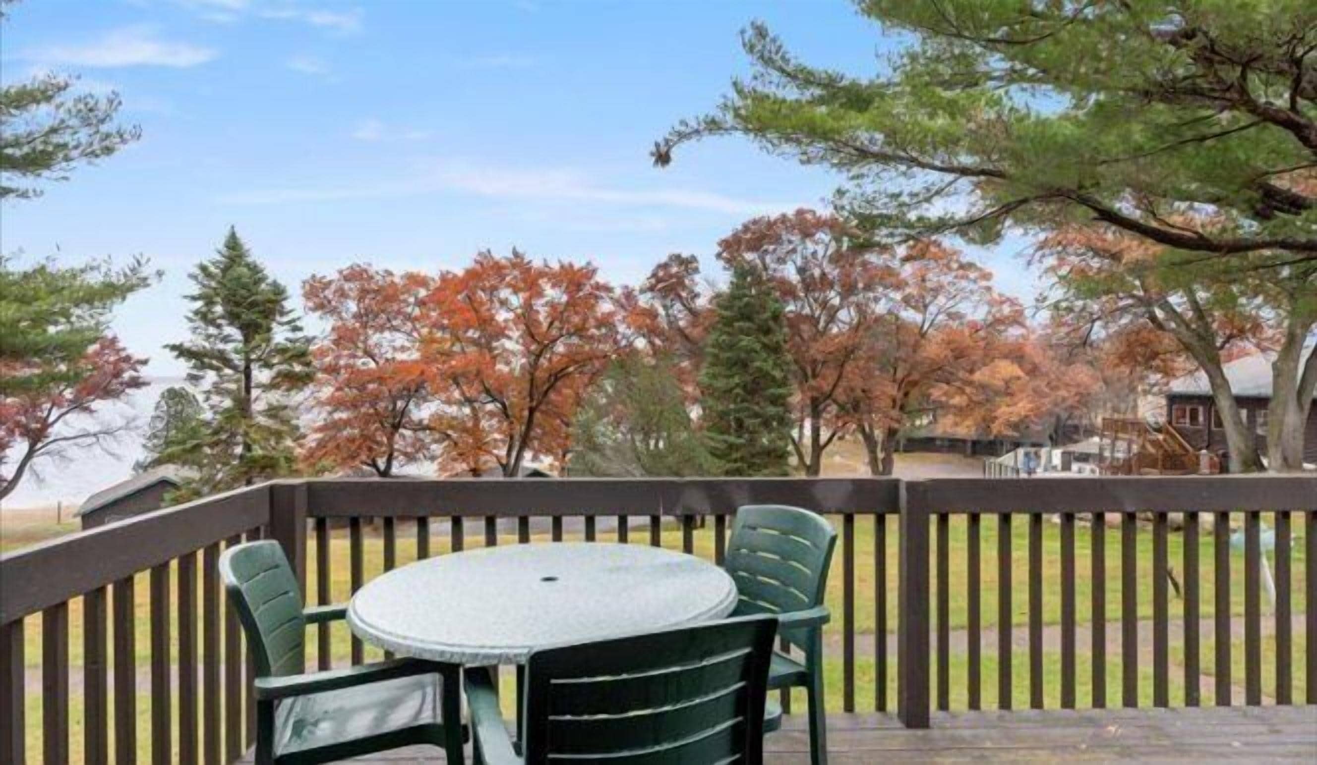 Une terrasse dotée d’une table ronde et de chaises, entourée d’une rambarde en bois, d’où l’on a vue sur un parc arboré et un bâtiment situé au loin.