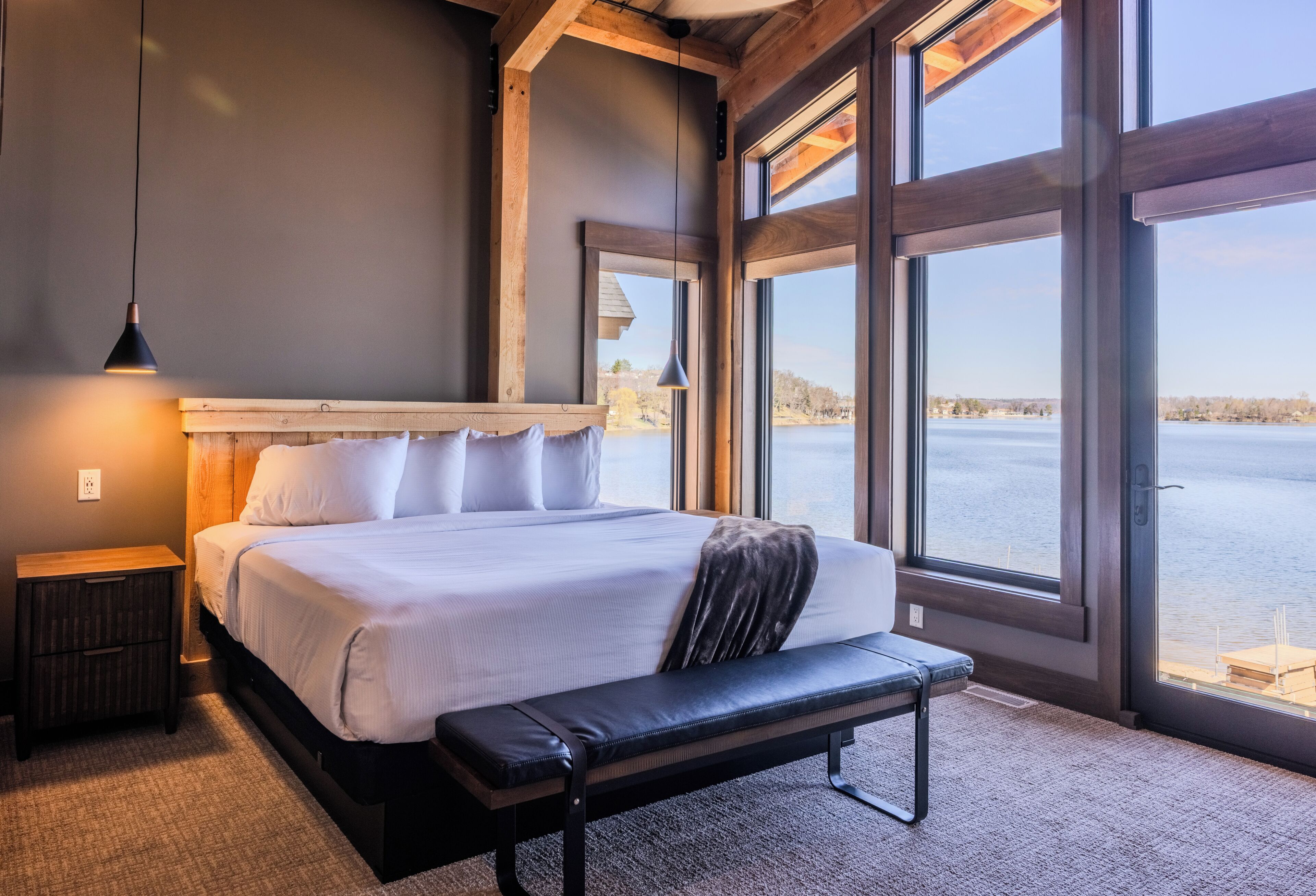 Una camera da letto con un letto grande, una panca, un comodino e vista su uno specchio d'acqua attraverso ampie finestre.