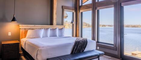 A bedroom with a large bed, a bench, a nightstand, and a view of a body of water through large windows.