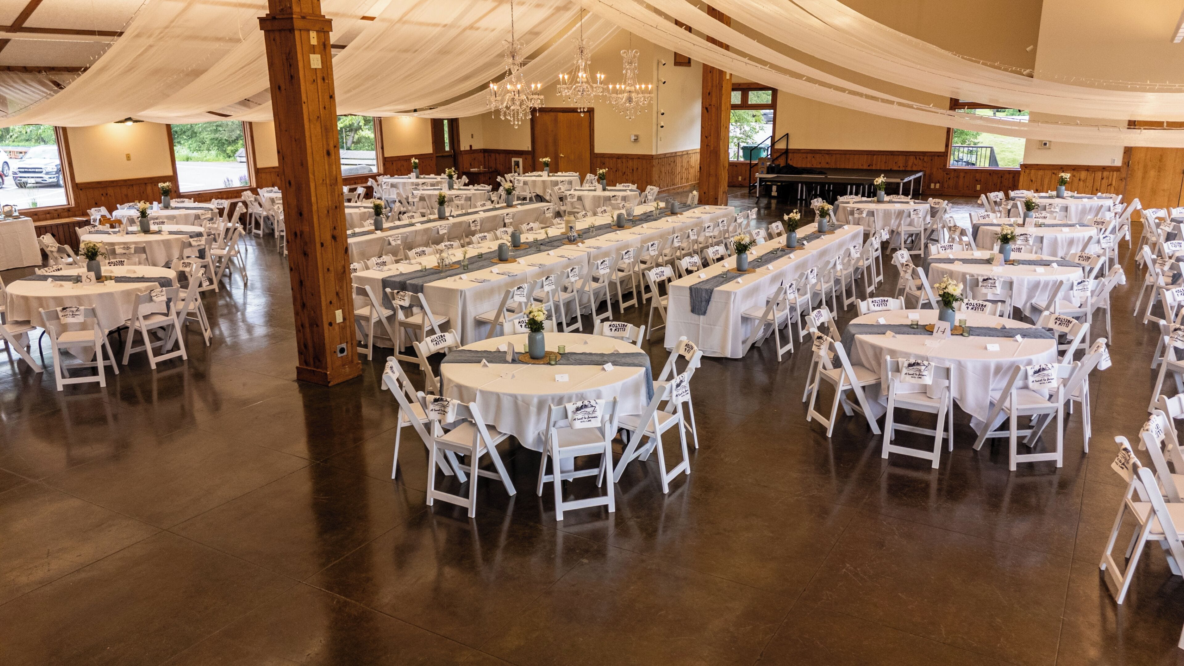 A banquet hall with multiple round tables, white chairs, and chandeliers.