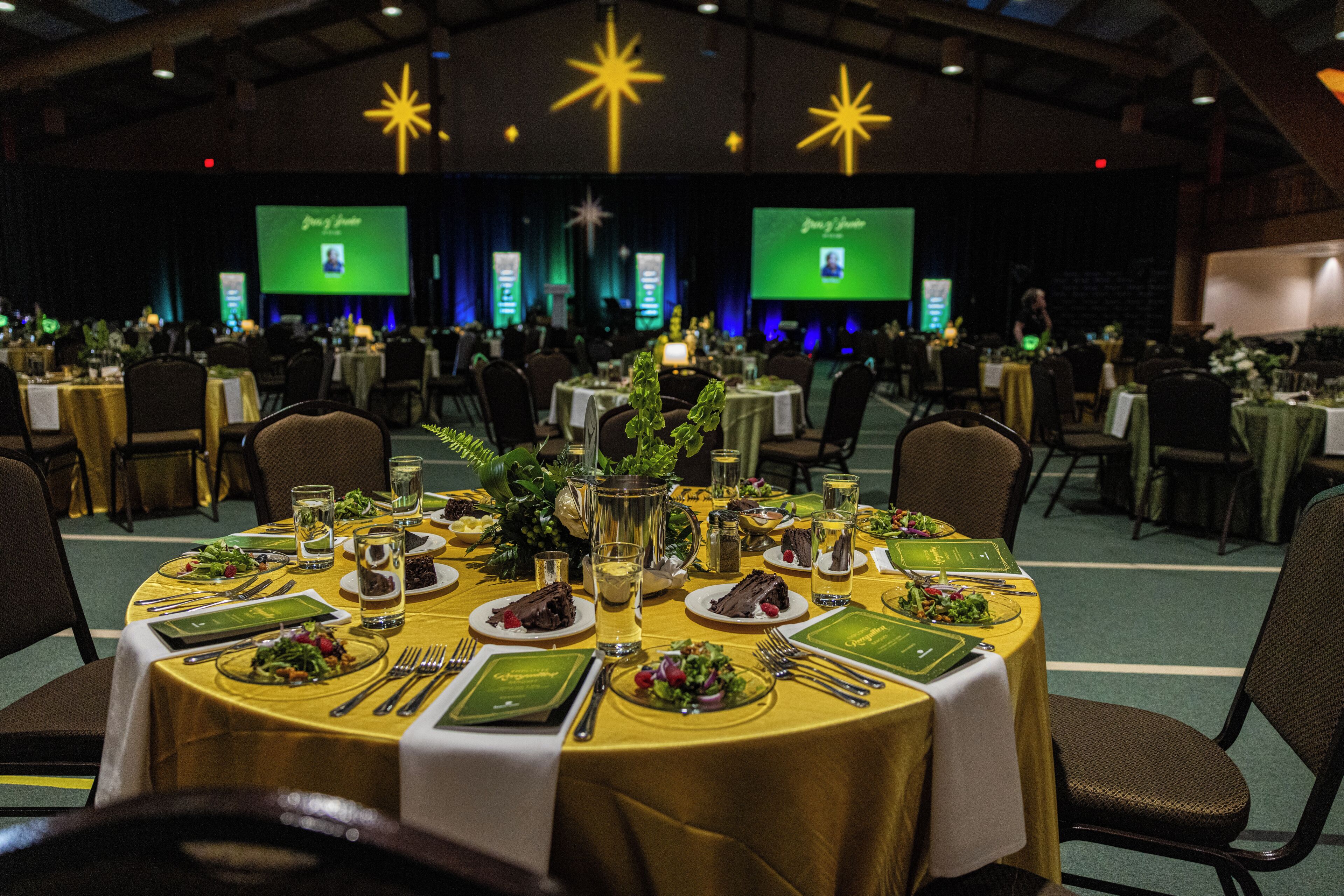 A banquet hall with round tables set for a formal event, featuring green menus, clear glassware, and floral centerpieces.