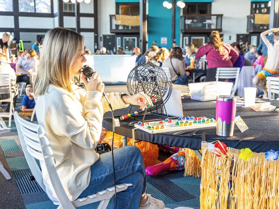 A person sitting at a table with a microphone and a game board, surrounded by other tables and chairs.