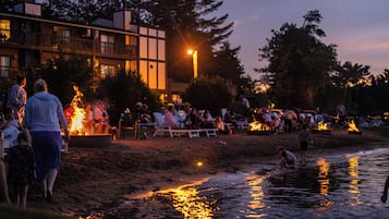 Reunión en la playa con gente alrededor de fogatas, un edificio al fondo y una masa de agua tranquila que refleja luces.